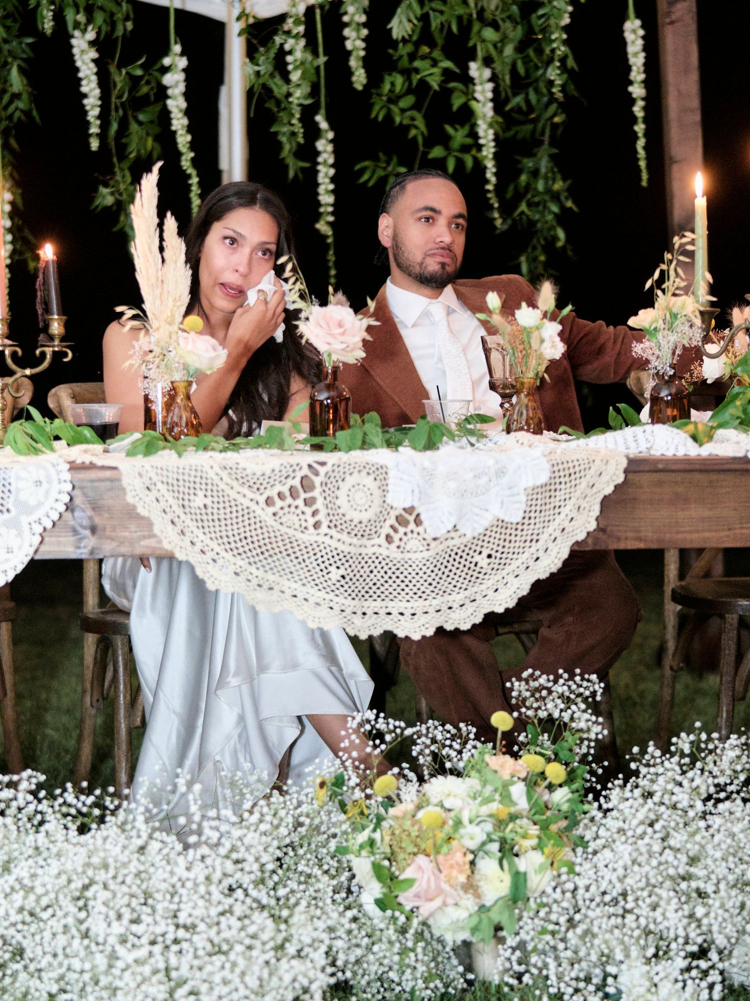 Wedding couple seated at decorated reception table with lace details and hanging floral arrangements.