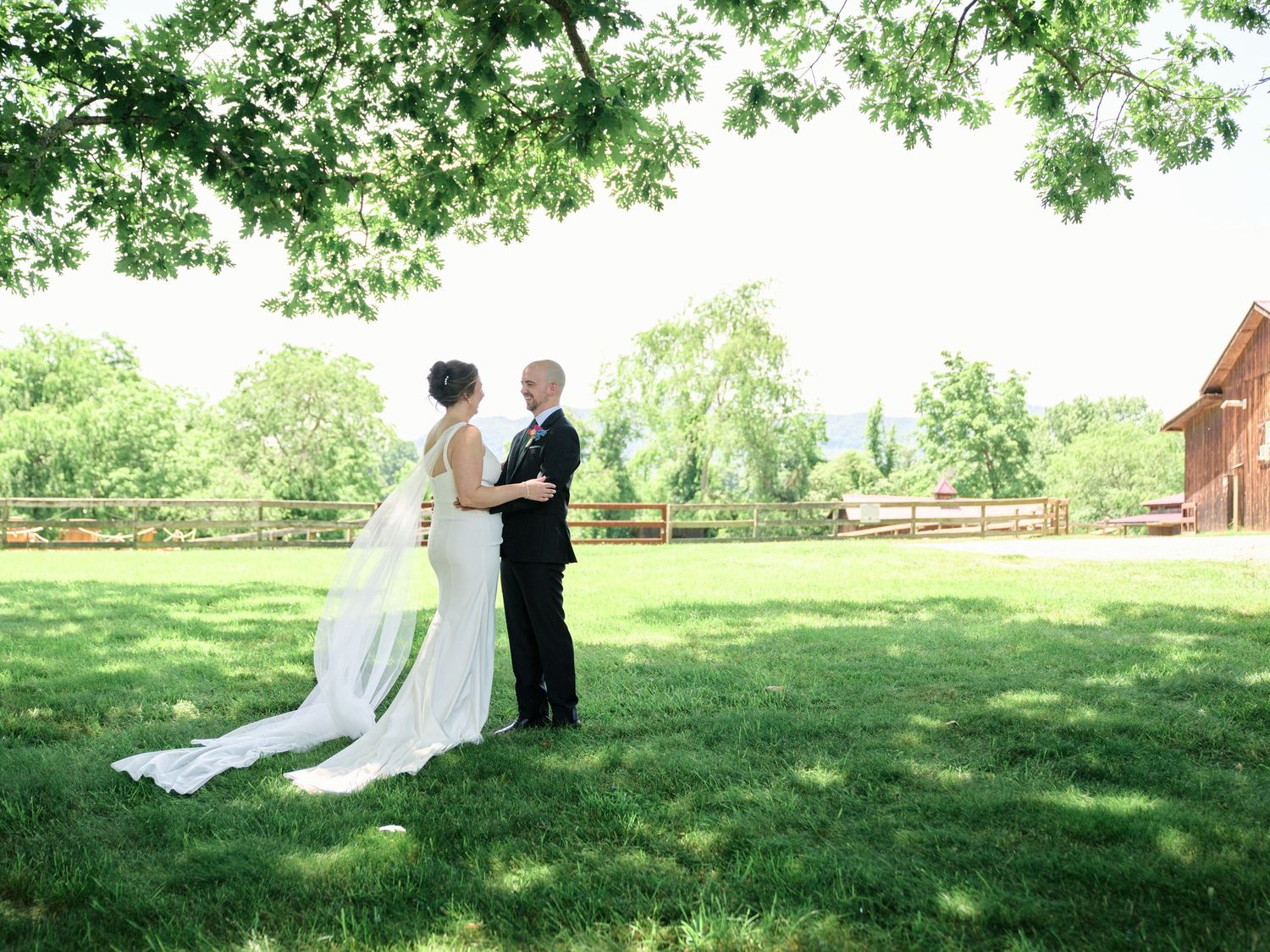 Wedding couple embraces on a sunny lawn with a rustic barn in the background.