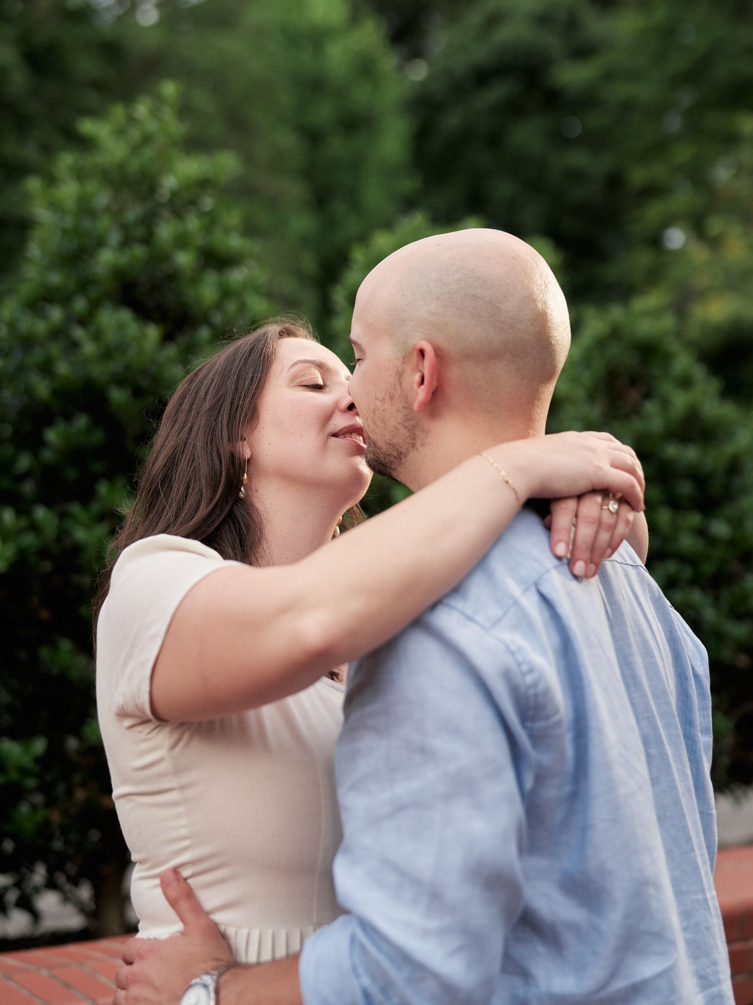 A romantic couple sharing a tender kiss in front of lush green bushes.