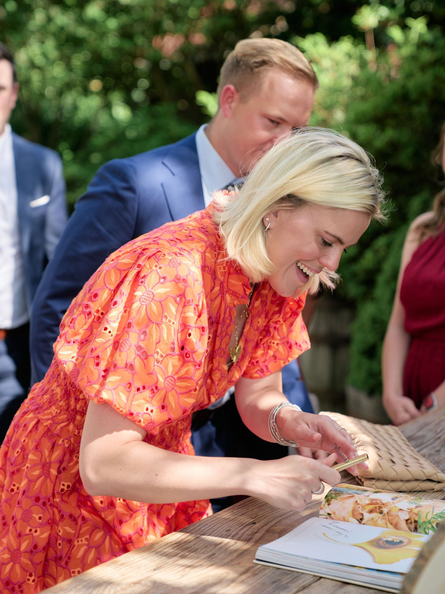 A guest in a bright coral dress signs the wedding guestbook at an outdoor table.
