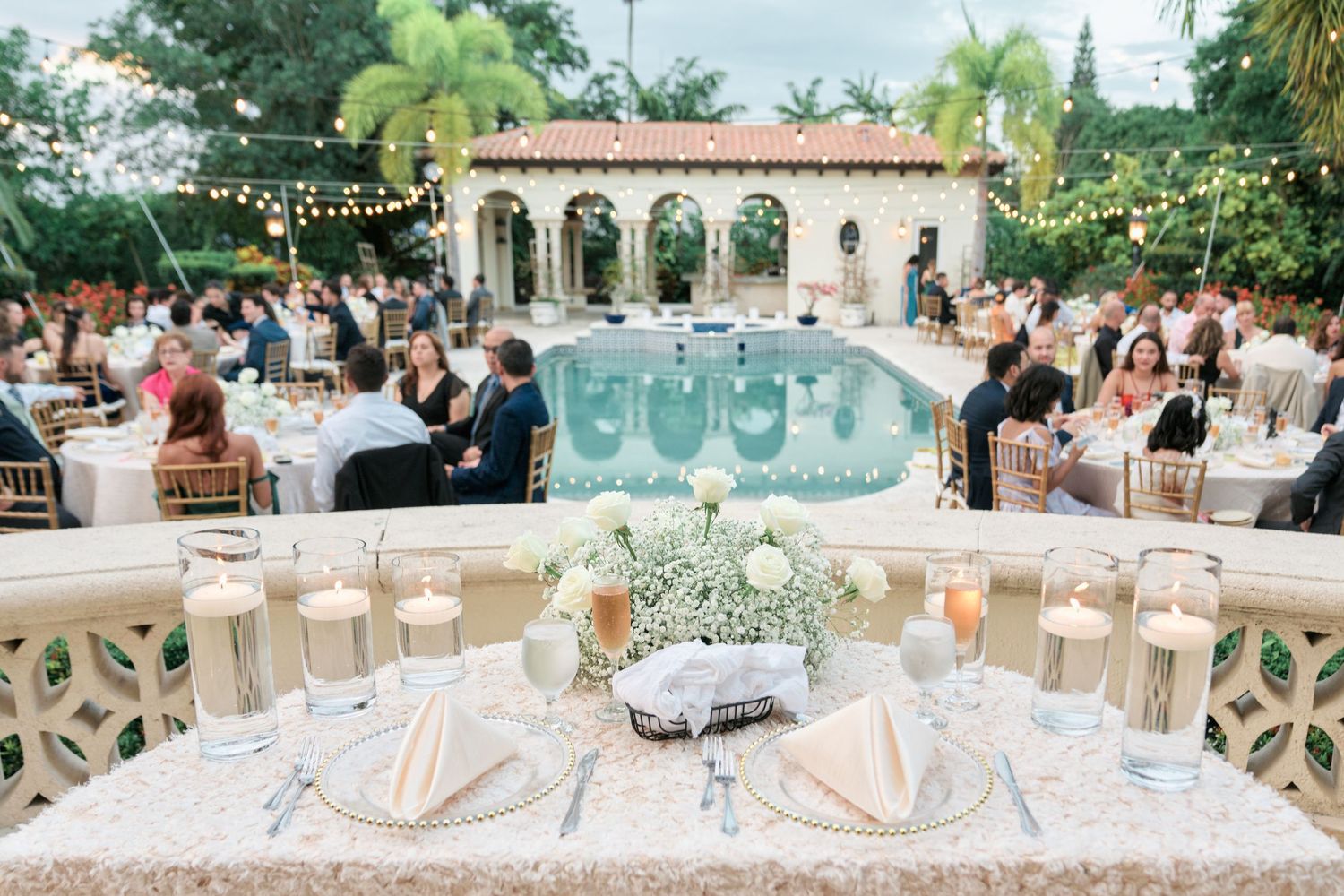 Elegant outdoor wedding reception setup with tables surrounding a pool and Mediterranean-style architecture in the background.