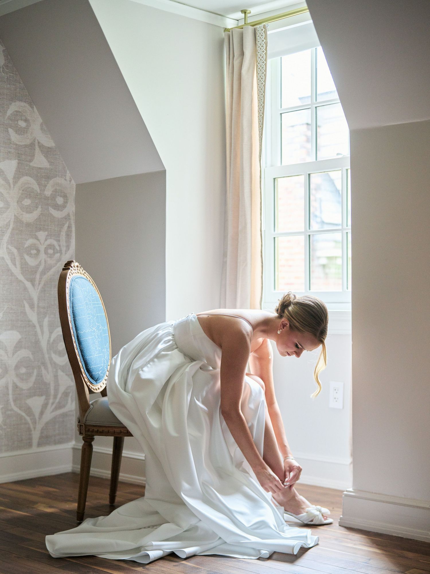 A person in a white wedding dress bends down to adjust their shoes by a window.