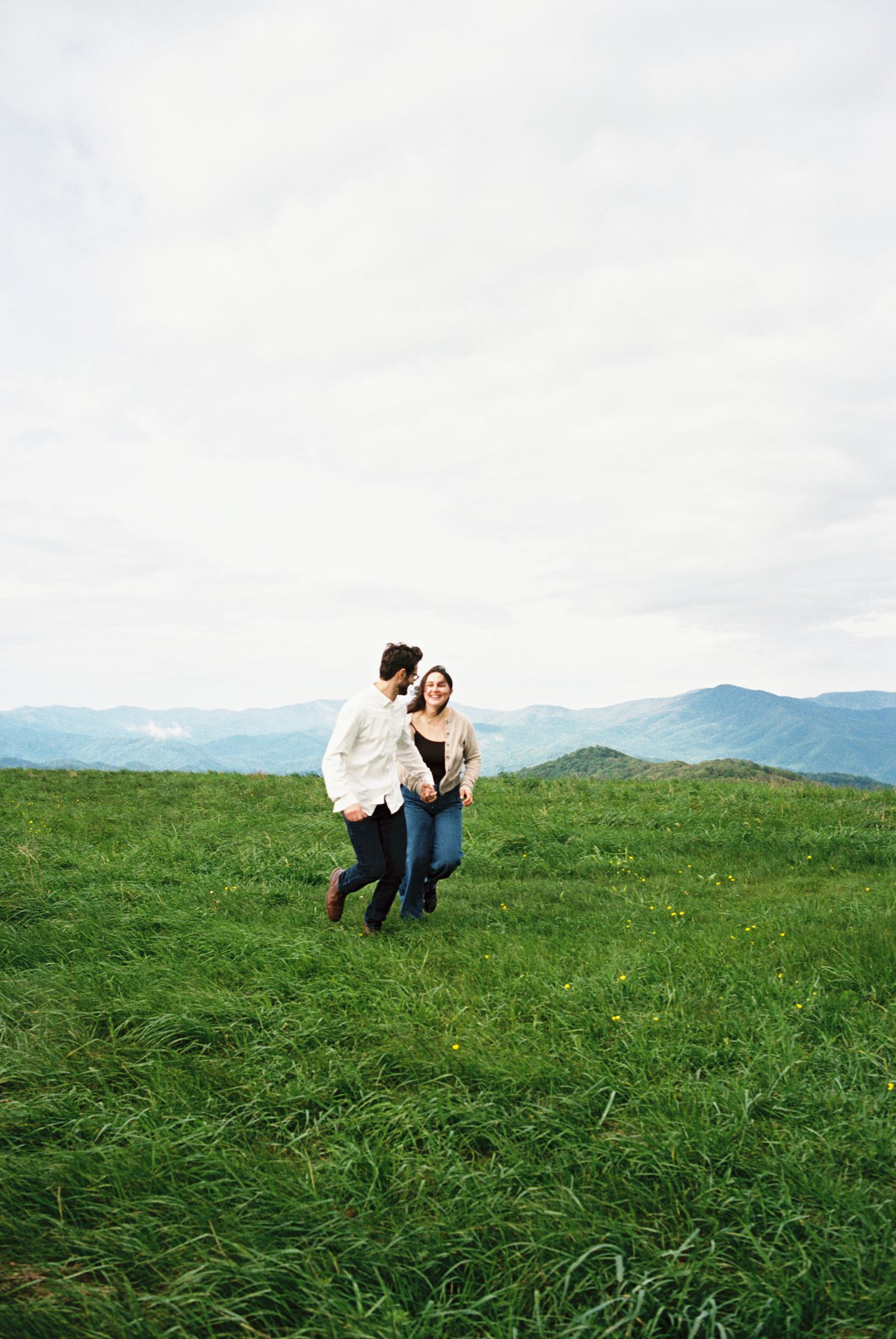 Couple frolicking on a lush green hilltop with distant mountains in the background under cloudy skies.