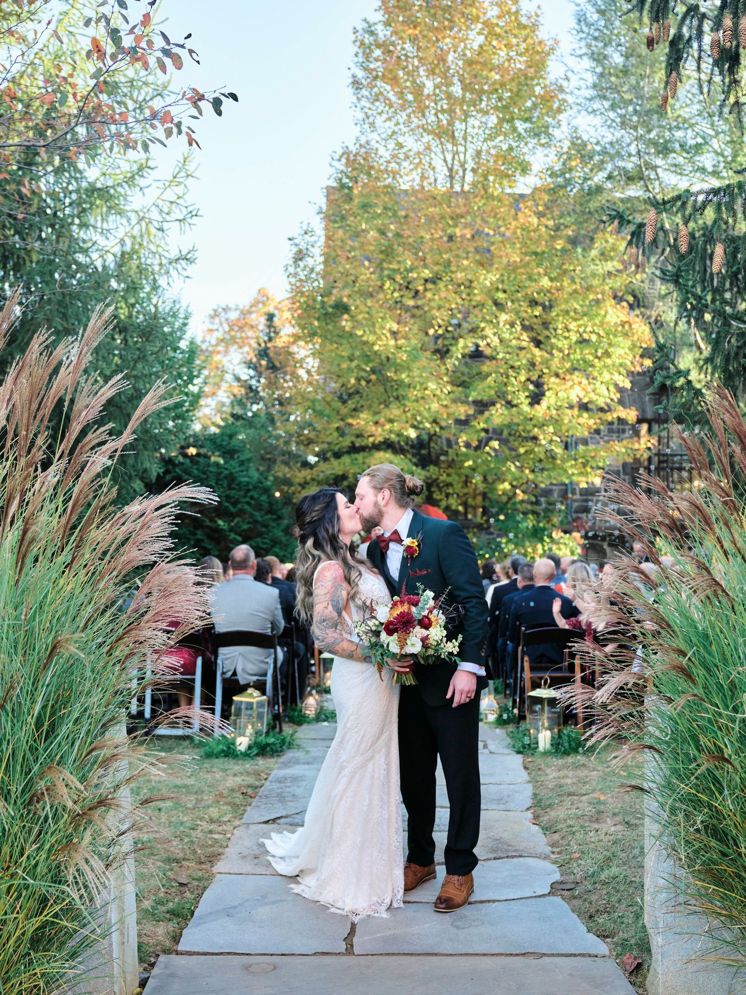 A couple walks down a pathway lined with ornamental grasses while wedding guests look on.
