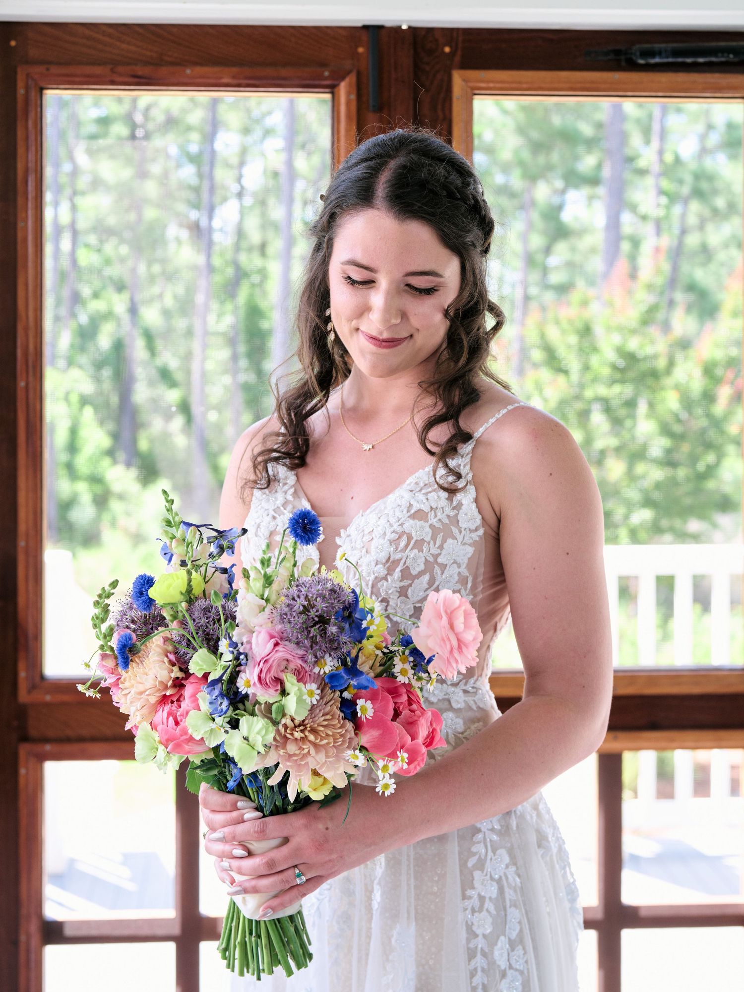 A bride in a lace wedding dress smiles while holding a vibrant bouquet of pink and blue flowers near sunny windows.