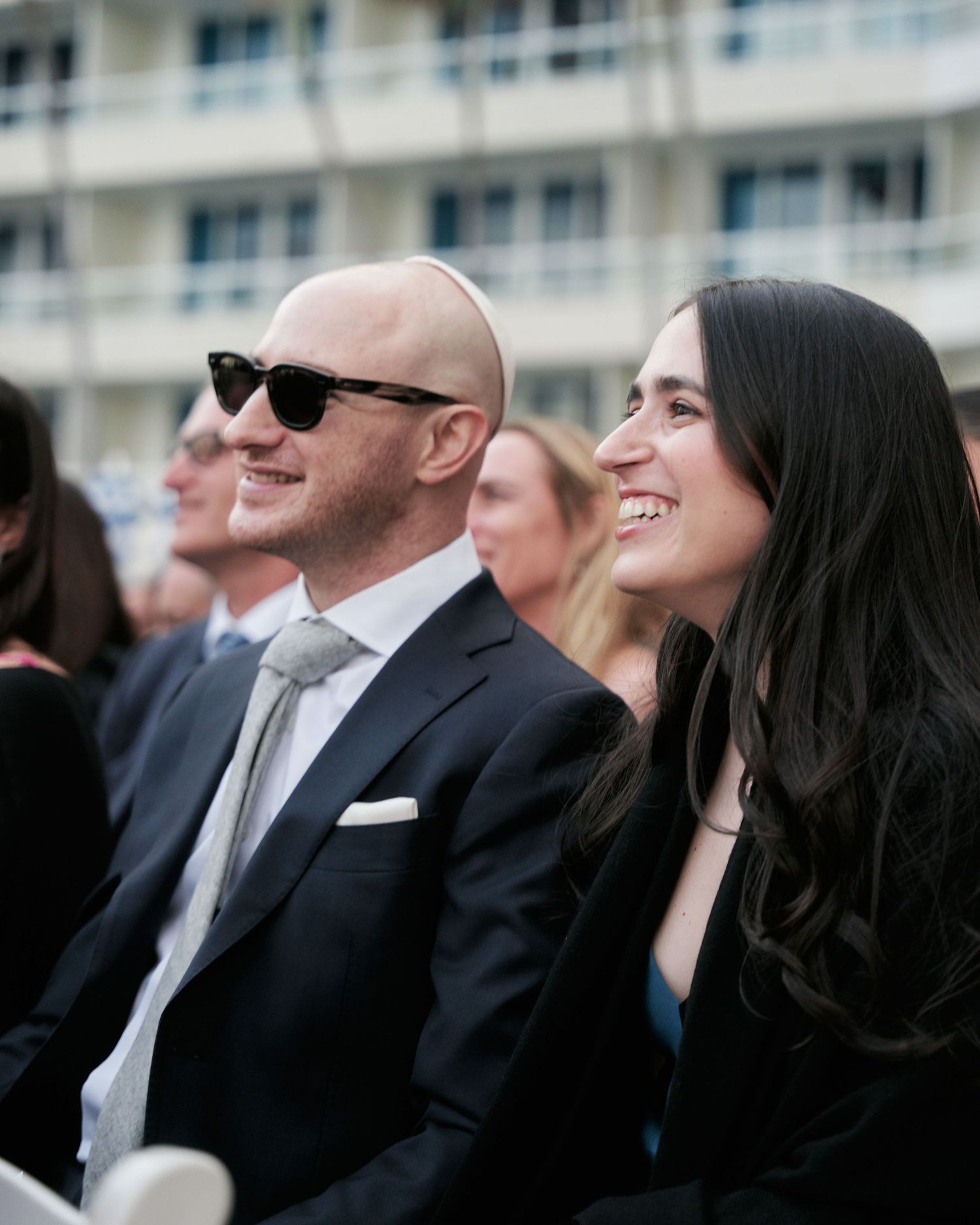 Wedding guests seated outdoors in formal attire smile and enjoy a sunny celebration in front of a modern building.