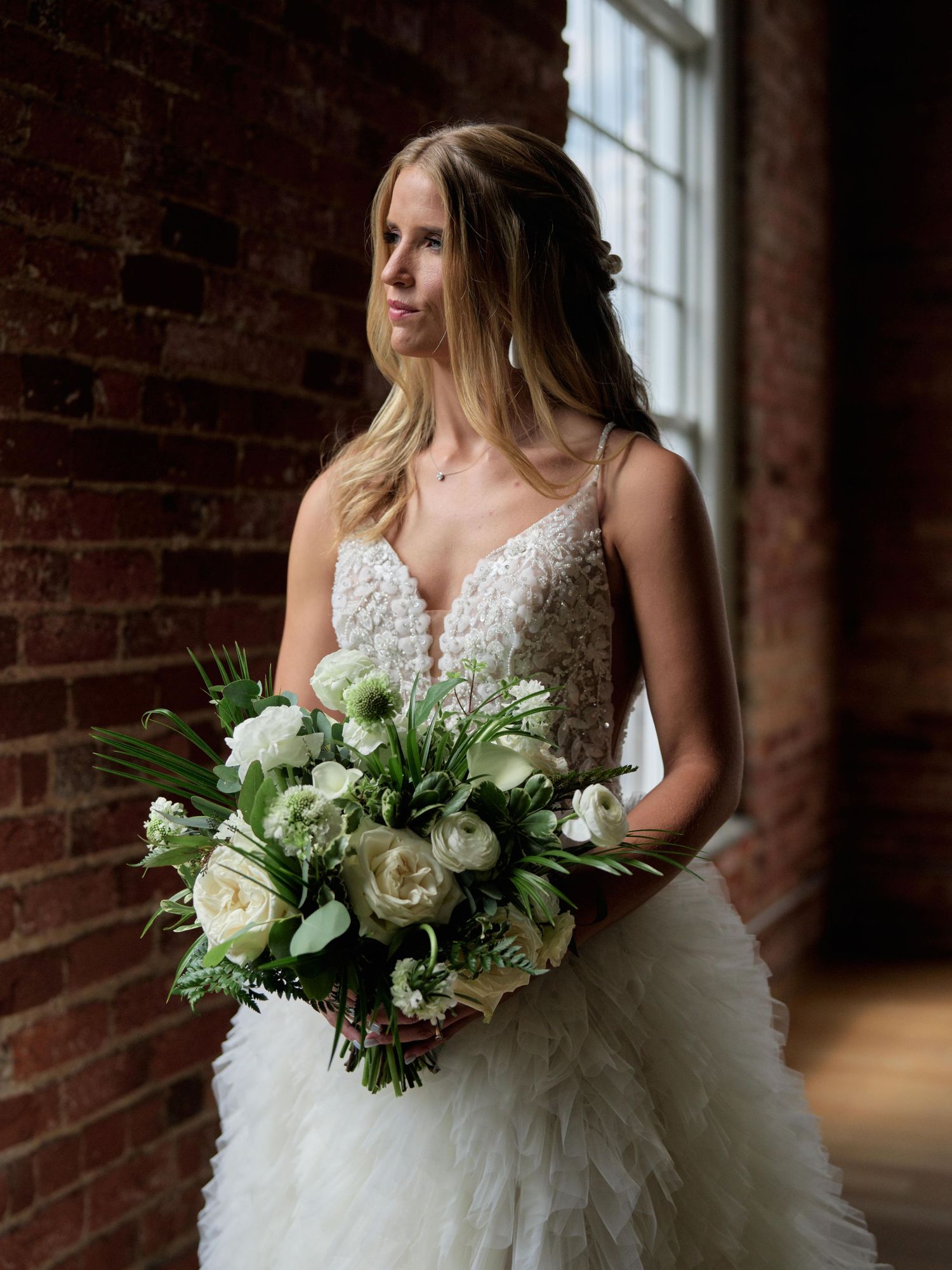Bride in lace wedding dress holds elegant white and green bouquet against rustic brick wall interior.