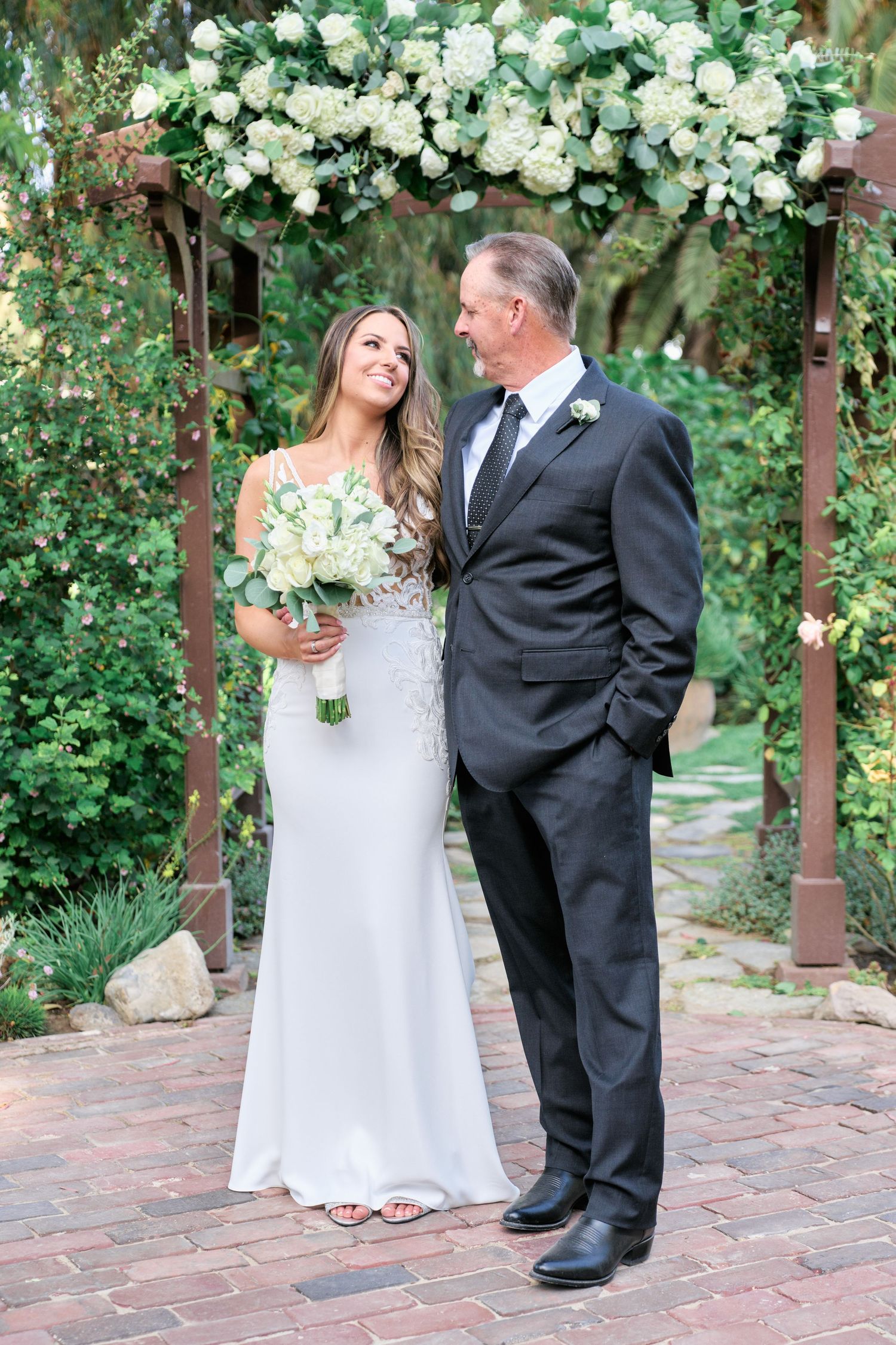 Elegant wedding portrait under a floral arch with white roses and lush garden backdrop.