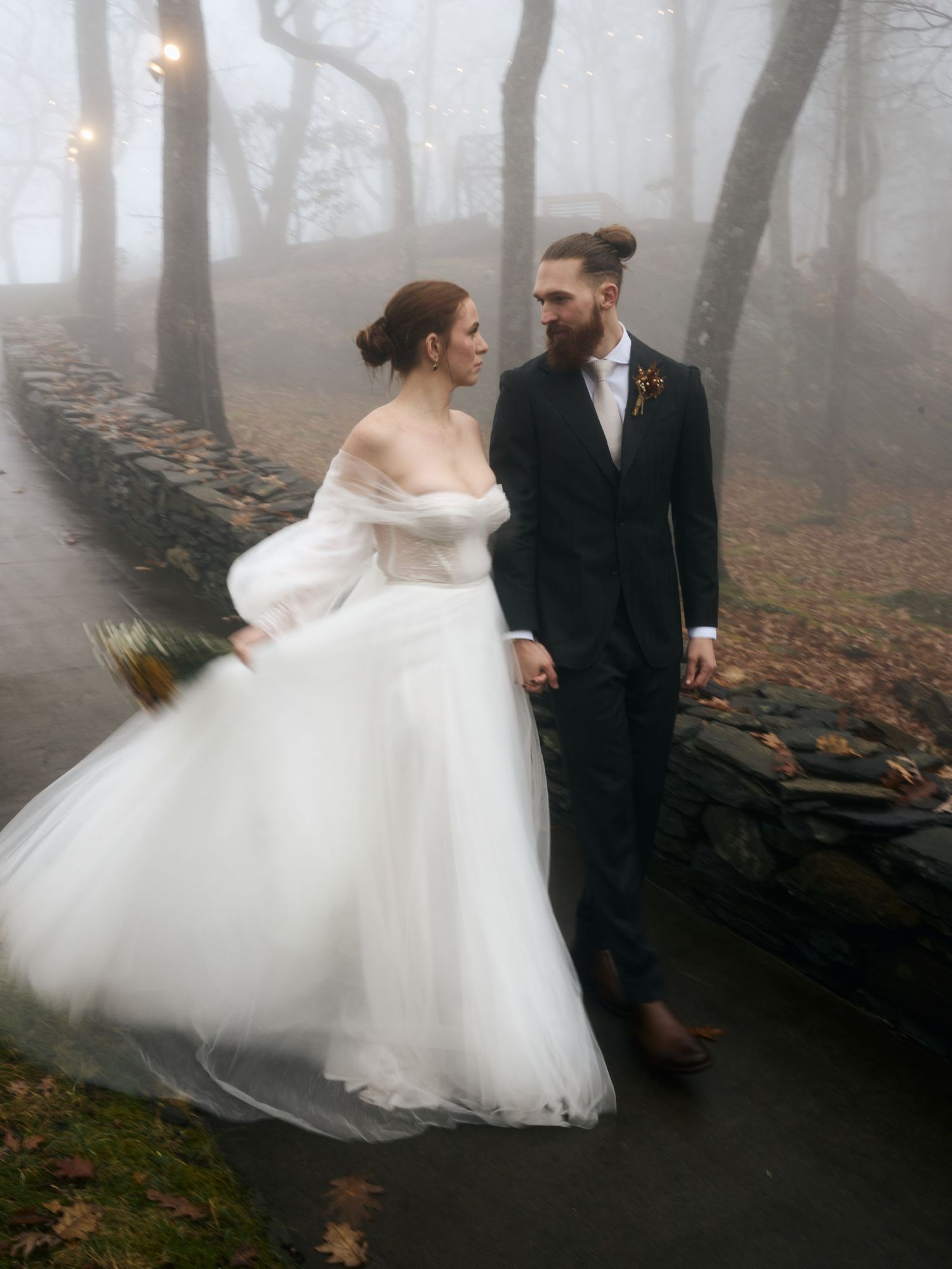 Moody wedding portrait on foggy path with illuminated trees and stone wall.