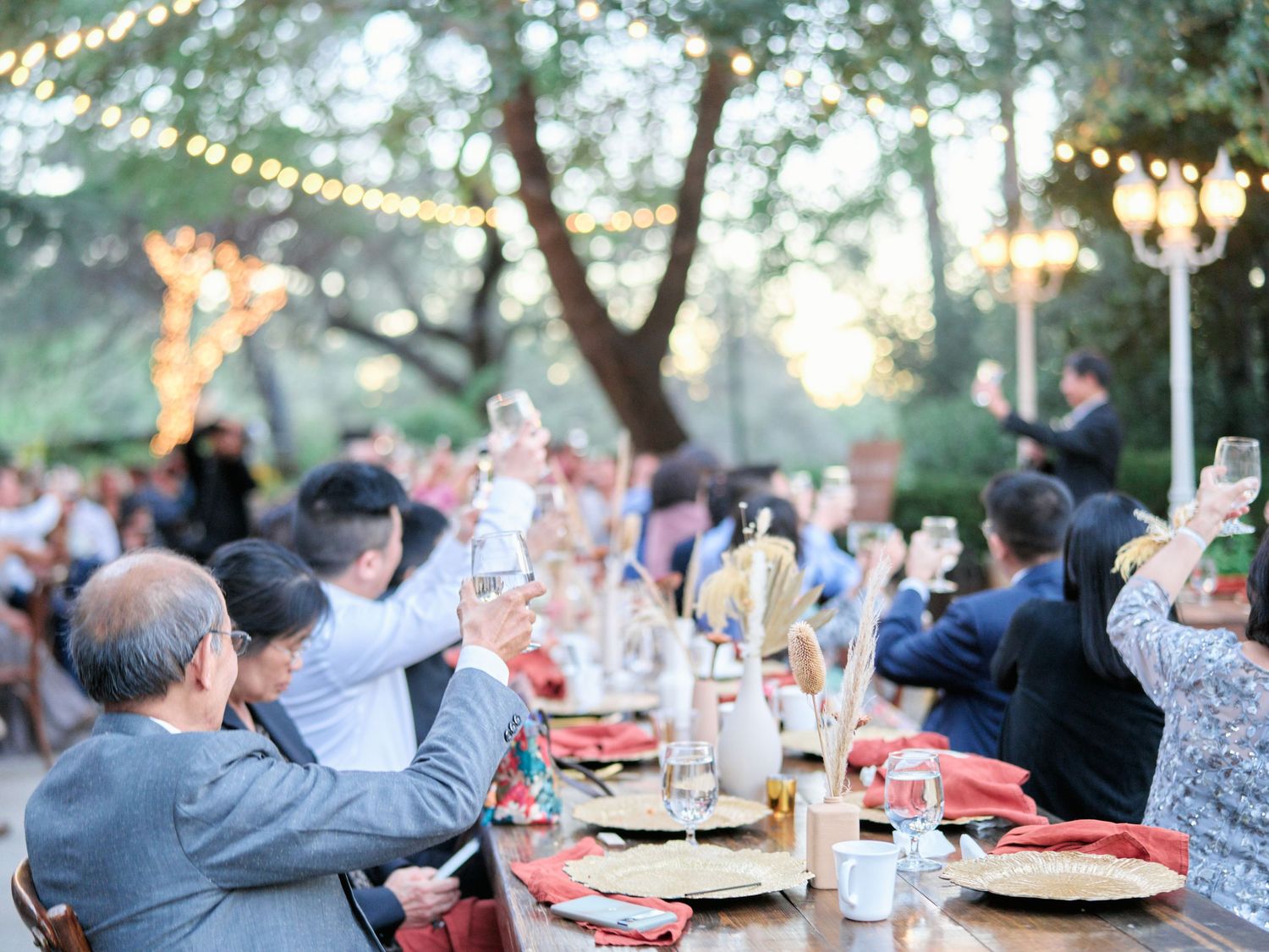 Wedding guests raise their glasses in celebration during an outdoor reception under string lights at sunset.
