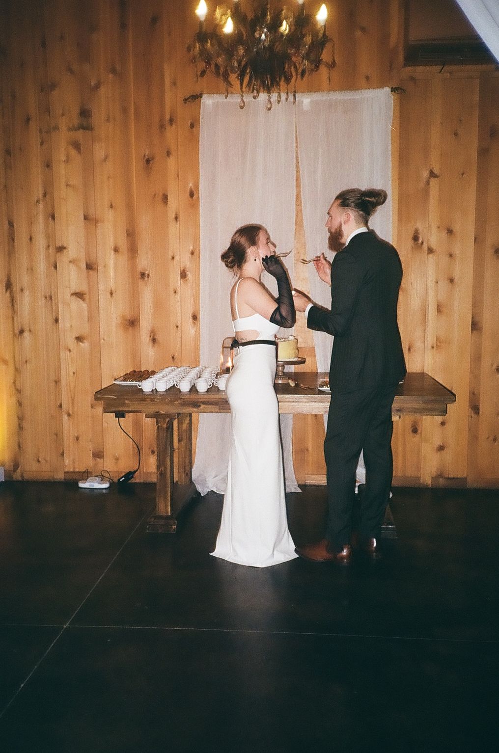 A couple feeds each other cake during their wedding reception in a rustic wooden venue with a chandelier.