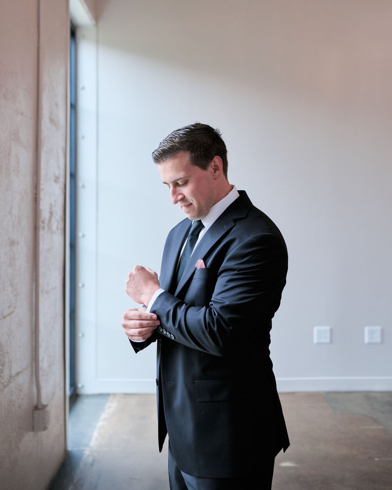 A groom in a dark suit adjusts his cufflinks while standing near a large window before his wedding ceremony.