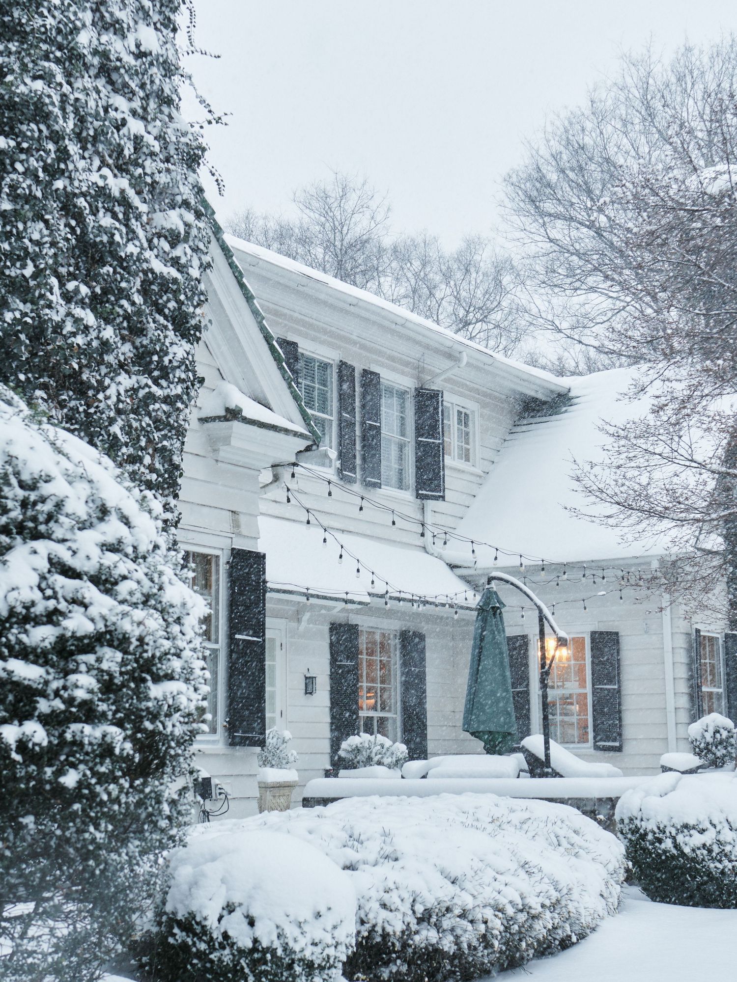 Snow-covered white colonial home with string lights, dark shutters, and frosted evergreen shrubs during a winter snowstorm.