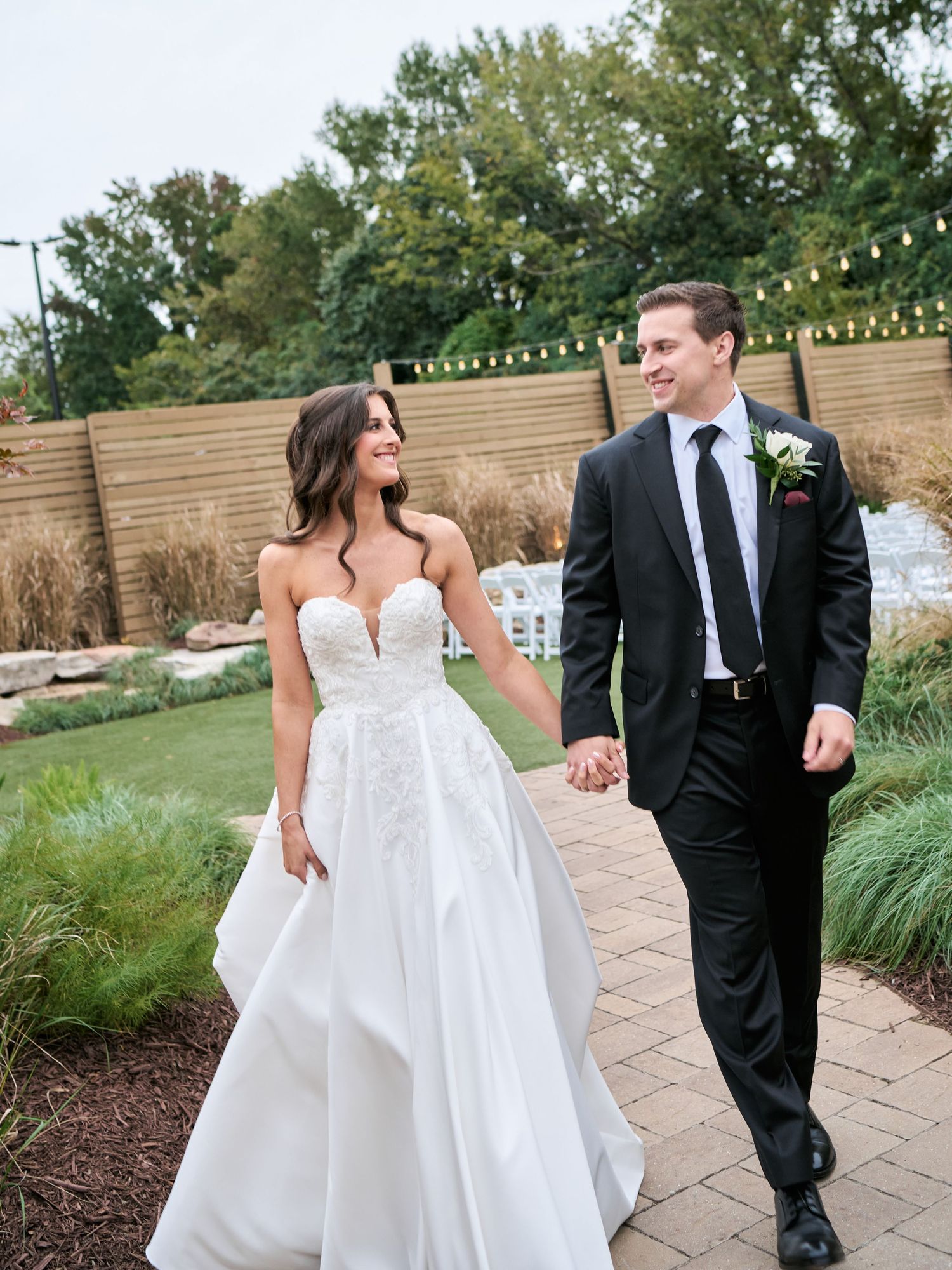 A couple in wedding attire walks together and shares joyful moments along a stone pathway in an outdoor garden setting.