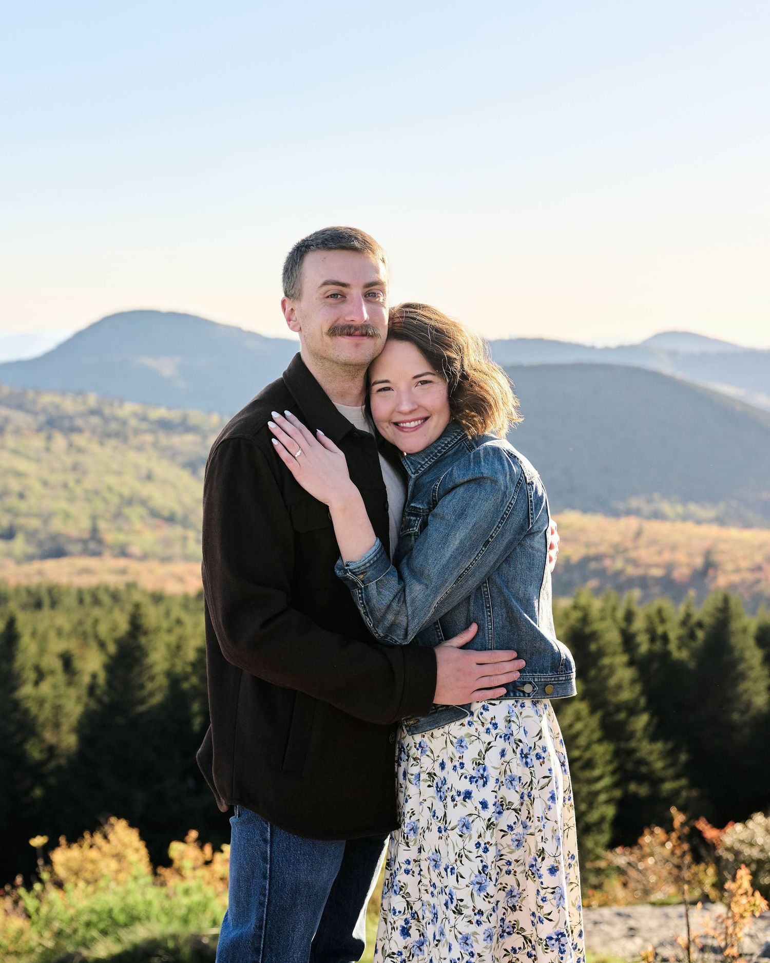 A couple embraces and smiles outdoors with a scenic mountain and evergreen forest backdrop on a clear day.