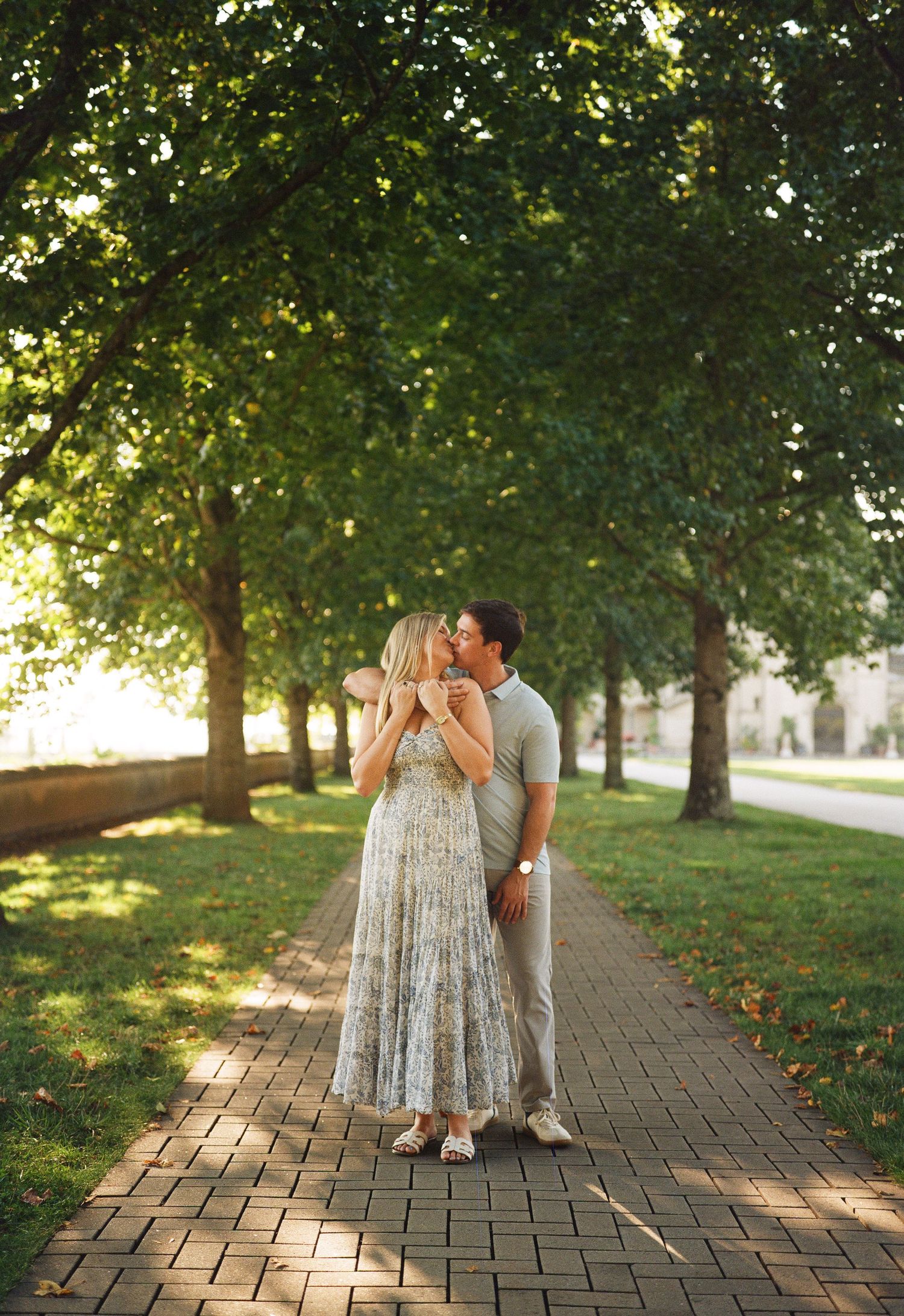 A romantic couple shares tender moments on a brick pathway surrounded by lush trees in golden sunlight.