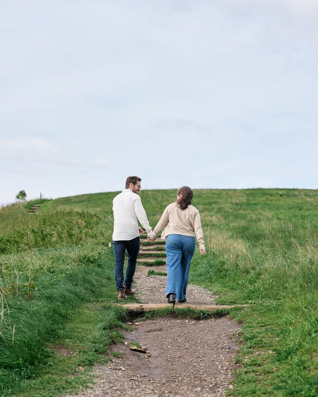 Two people walk up a dirt trail through rolling green hillside on a peaceful afternoon.