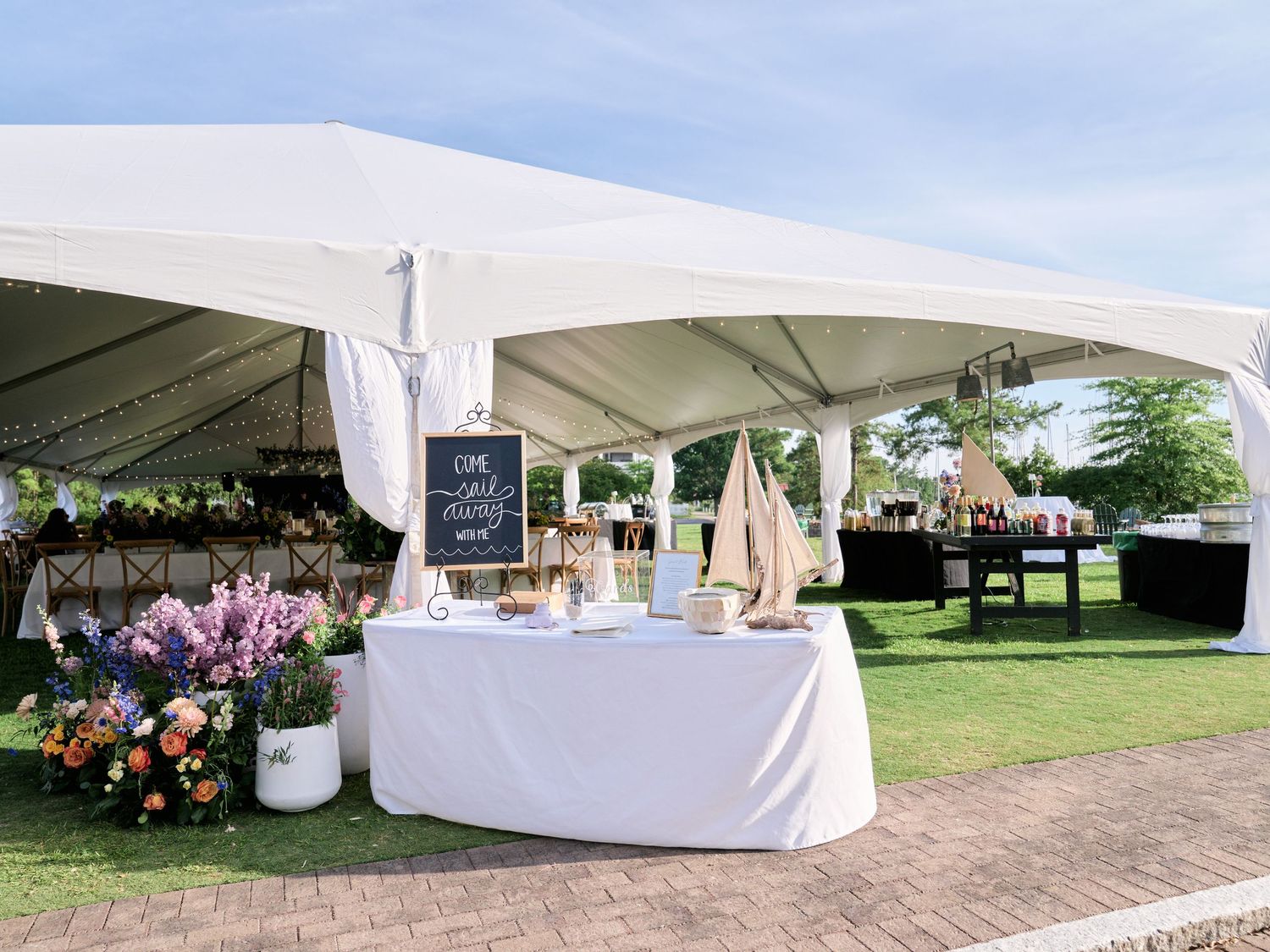 An elegant white wedding tent setup with decorative chalkboard signs, floral arrangements and sailboat accents on display.