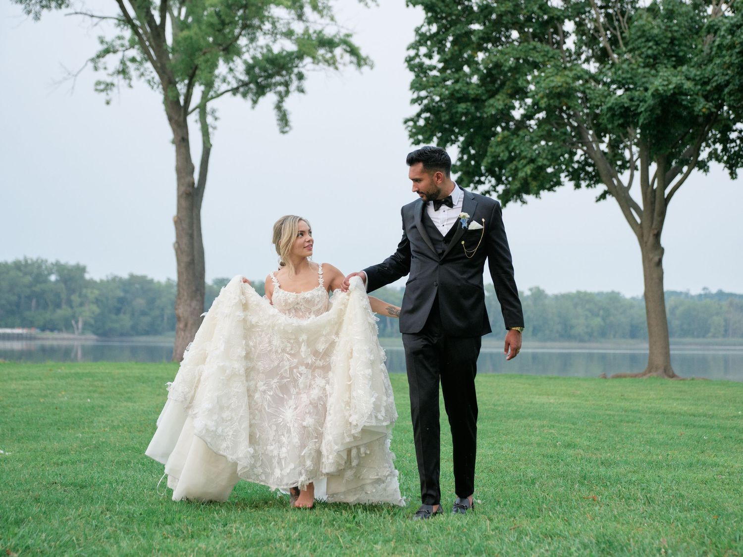 A couple in wedding attire walks together on a grassy lawn with trees and a lake in the background.