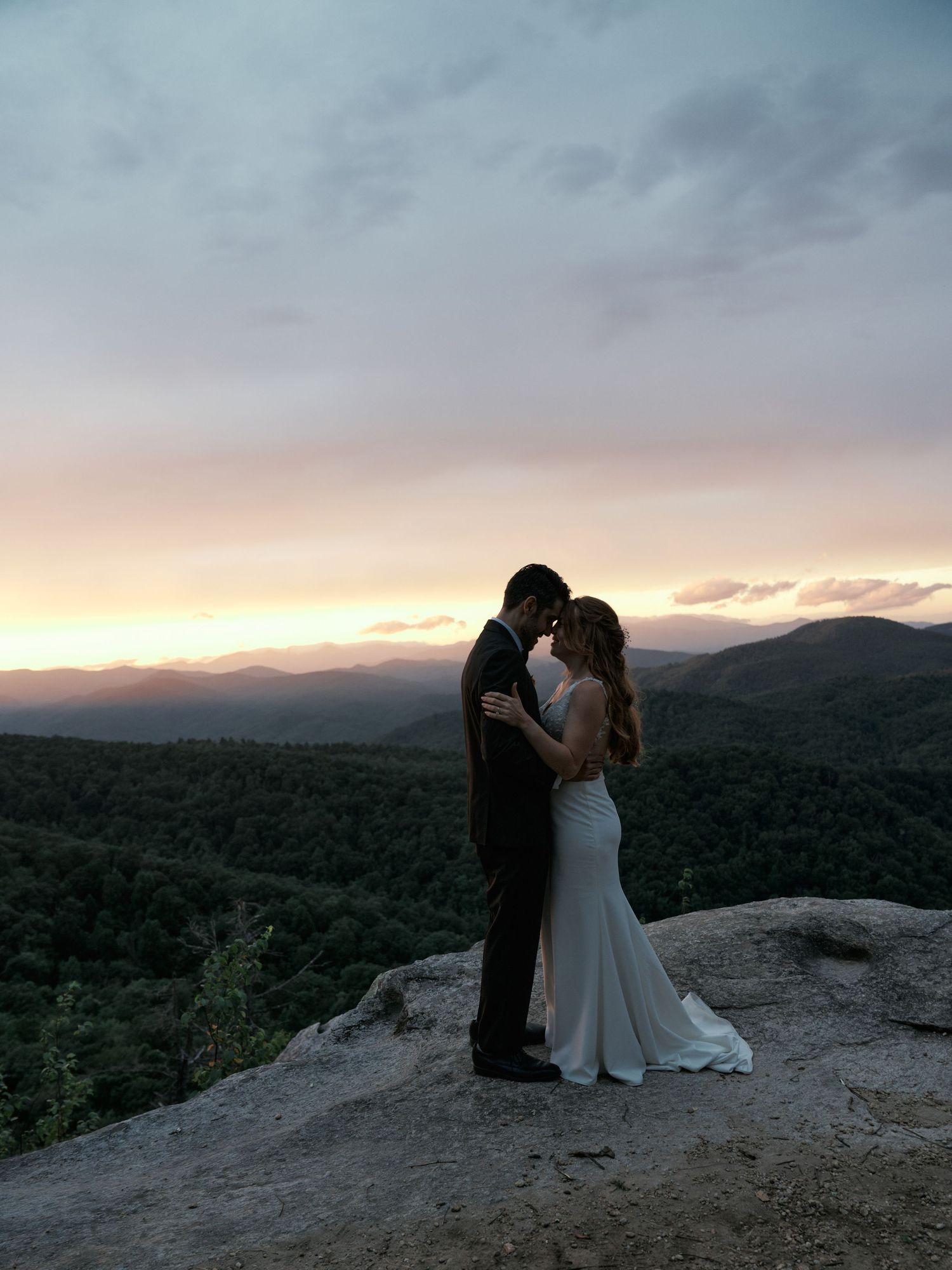 A romantic sunset silhouette of newlyweds embracing on a mountain overlook with vast mountain ranges behind them.