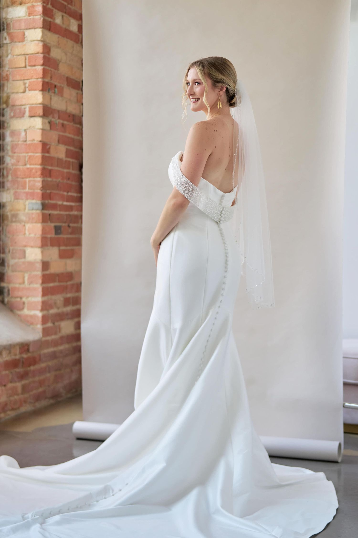 Elegant white wedding dress with button details and flowing train shown from multiple angles in a brick-walled room.