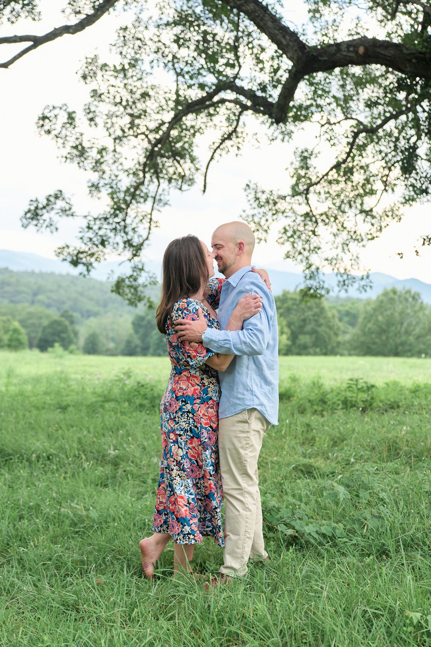 A couple embraces in a meadow under a large tree with mountains in the background.