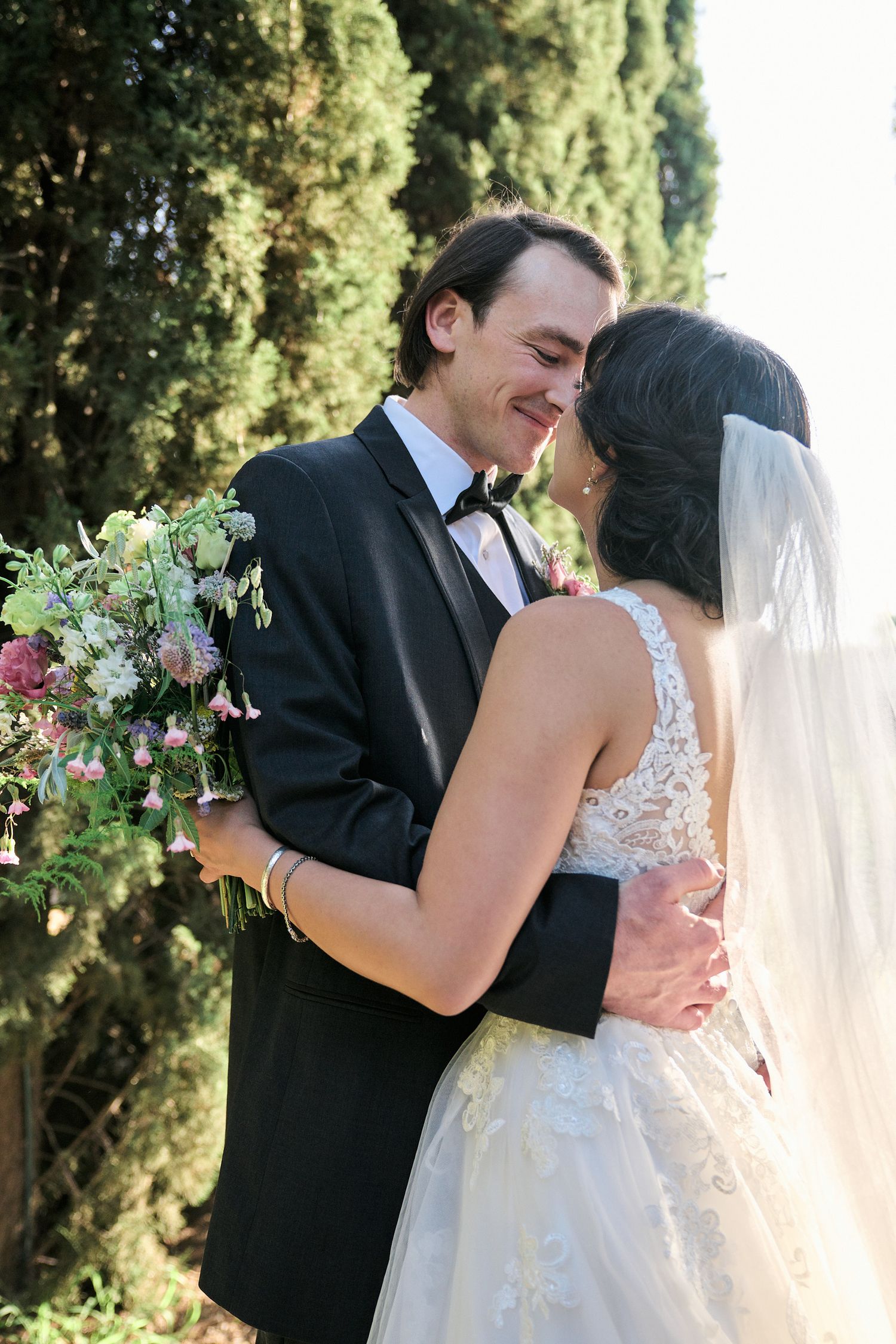 A romantic outdoor wedding moment captured among cypress trees with floral bouquet.