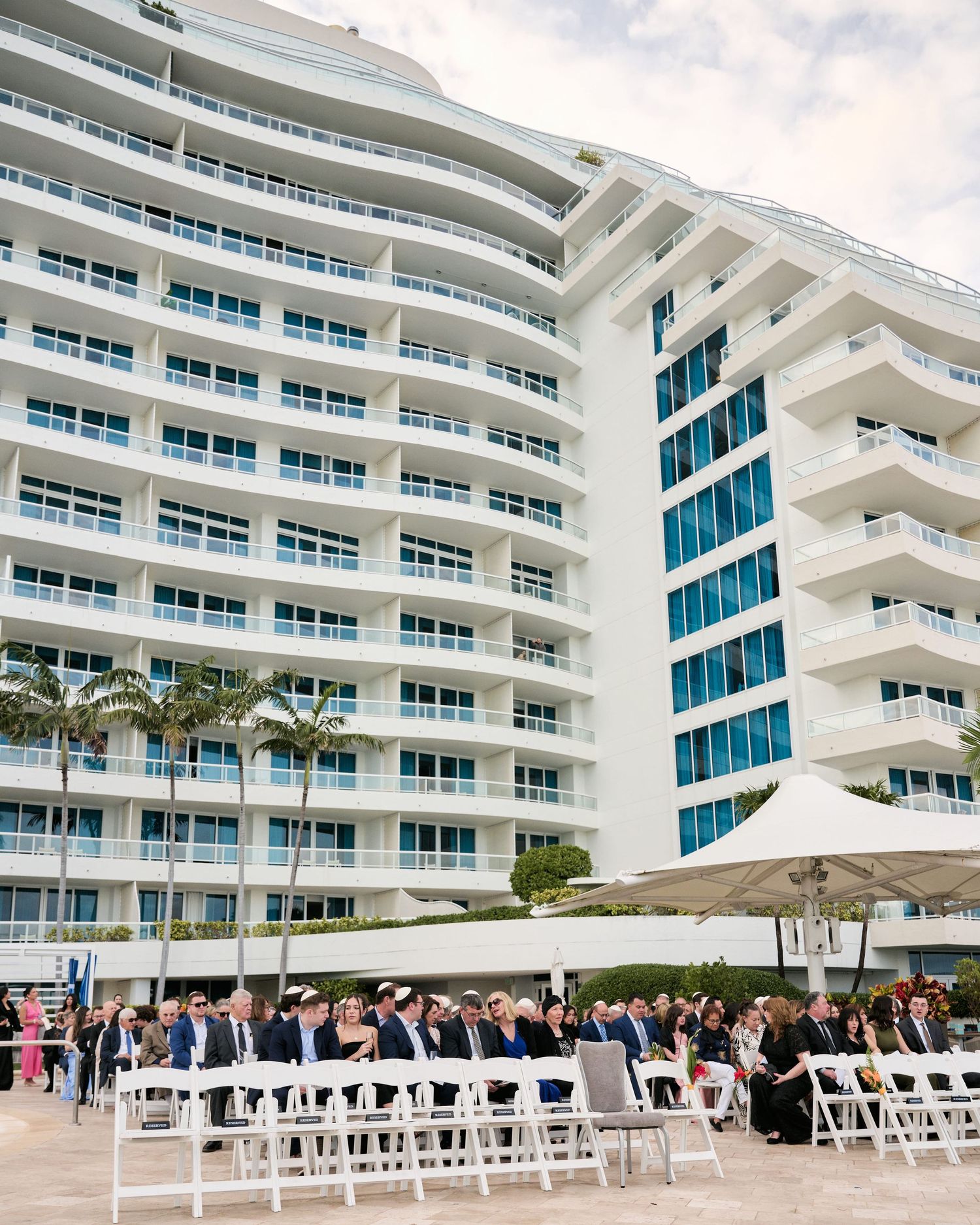 Modern white beachfront hotel building with rows of white chairs set up for an outdoor ceremony.