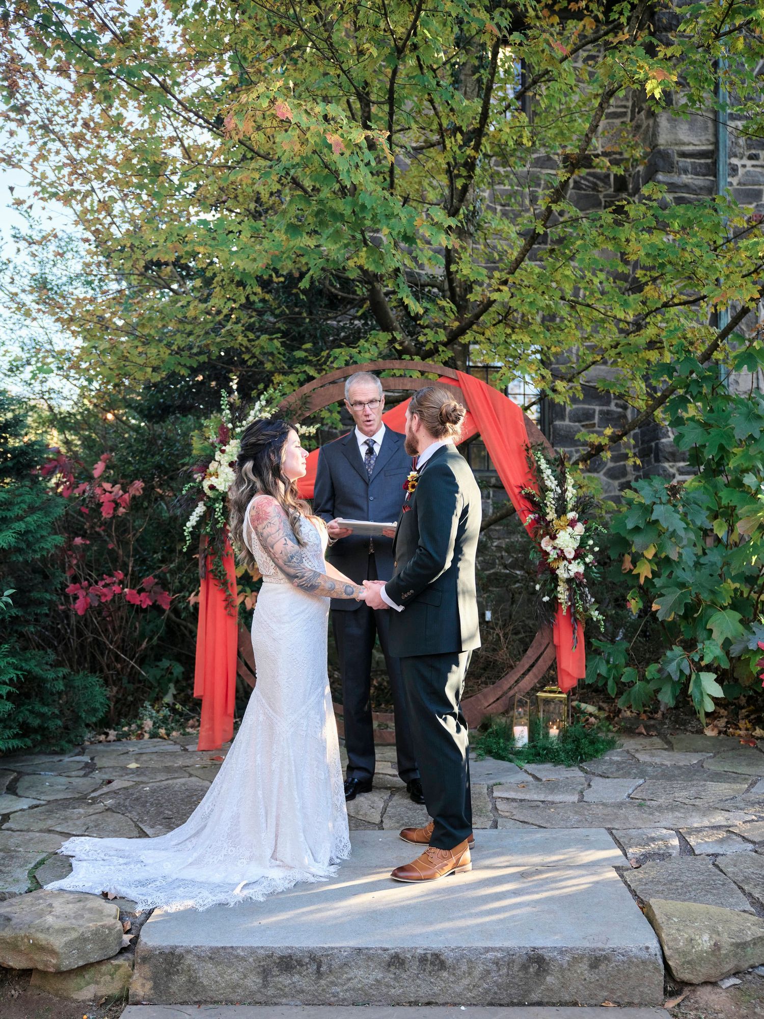 Wedding ceremony under a circular red arch decorated with white flowers on a stone patio amid autumn foliage.
