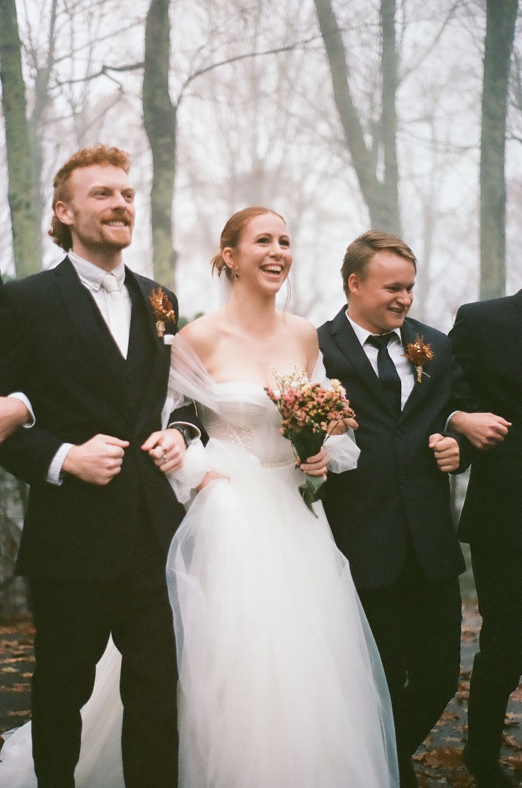 Wedding party laughing together on misty forest path covered in fallen leaves.