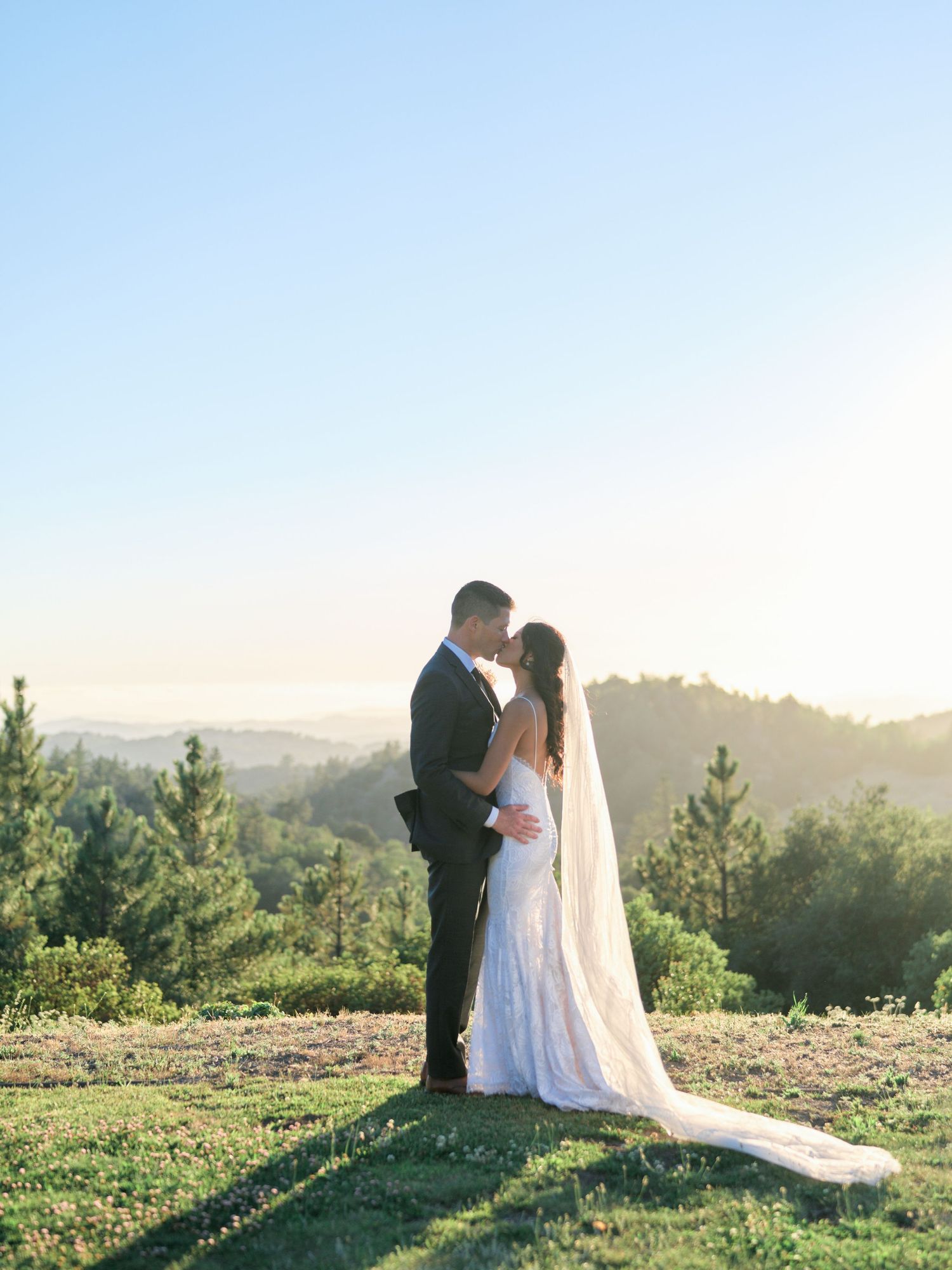 A romantic wedding portrait captured on a hilltop overlooking a mountain landscape at sunset.