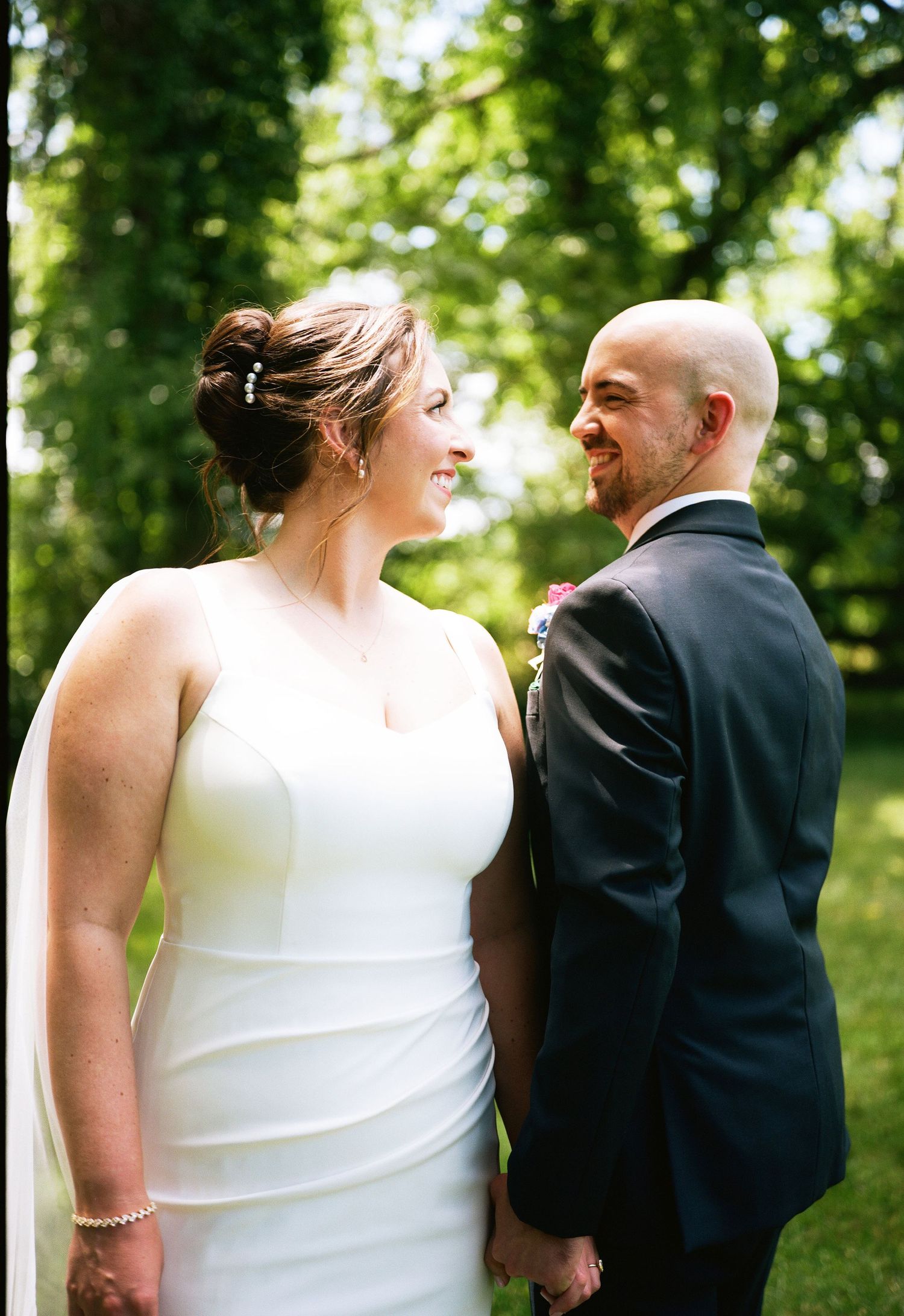 Smiling bride in white gown and groom in navy suit hold hands outdoors among lush green trees on their wedding day.