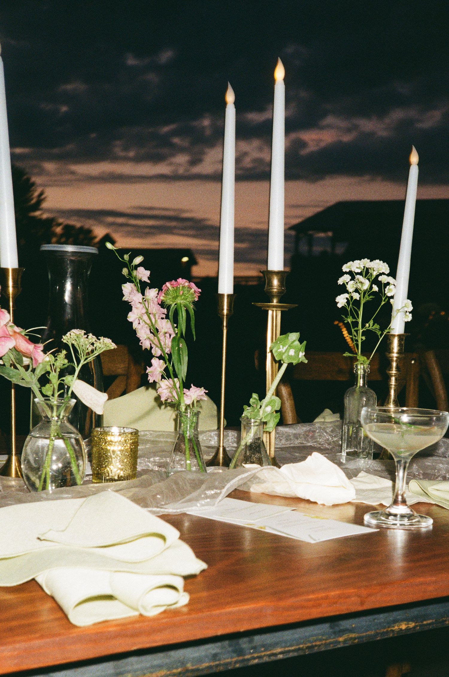 Elegant outdoor wedding table with tall brass candlesticks, small floral vases, and a martini glass at sunset.