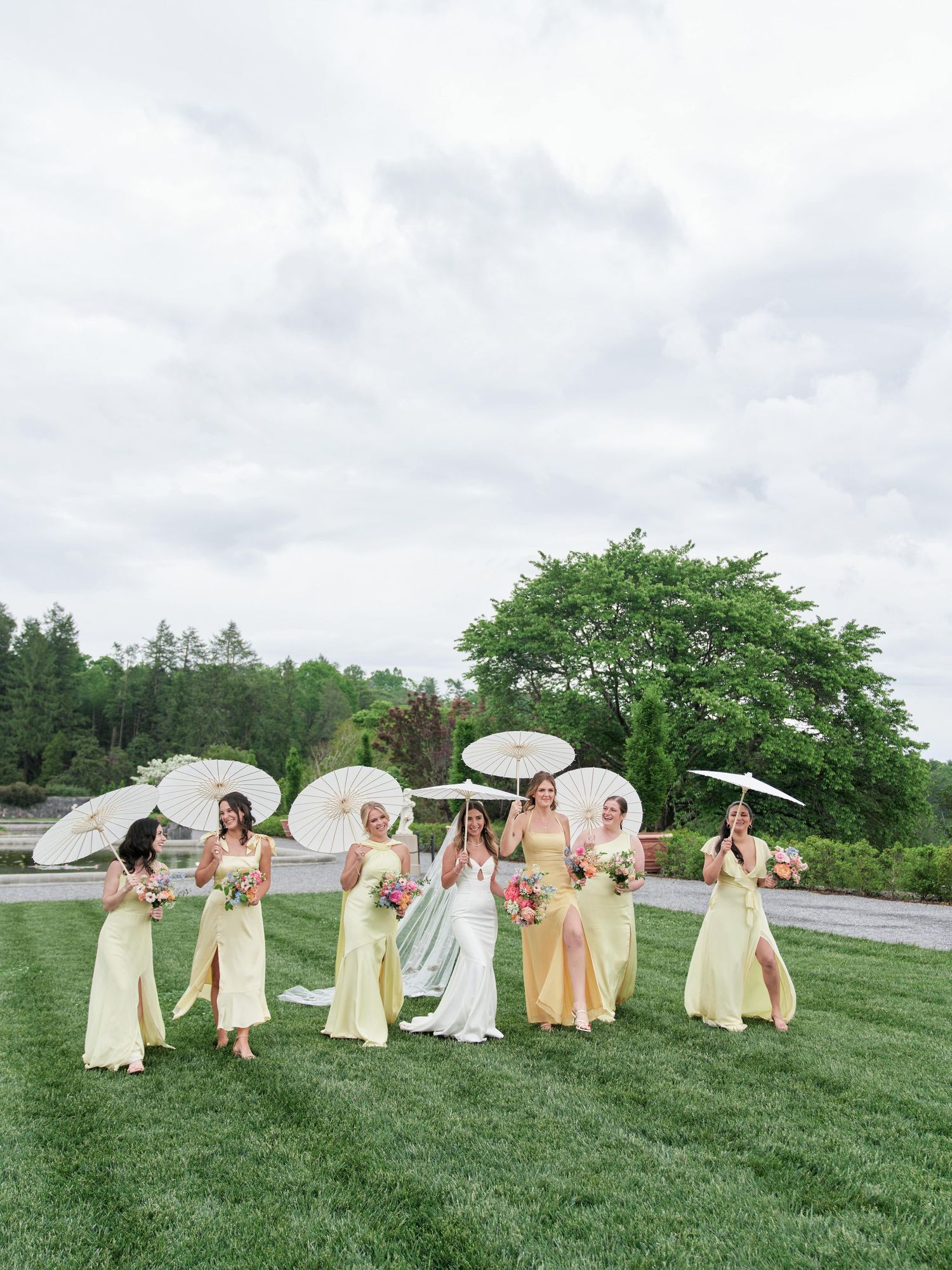 Wedding party in pastel yellow dresses holding white parasols pose on green lawn under cloudy sky.