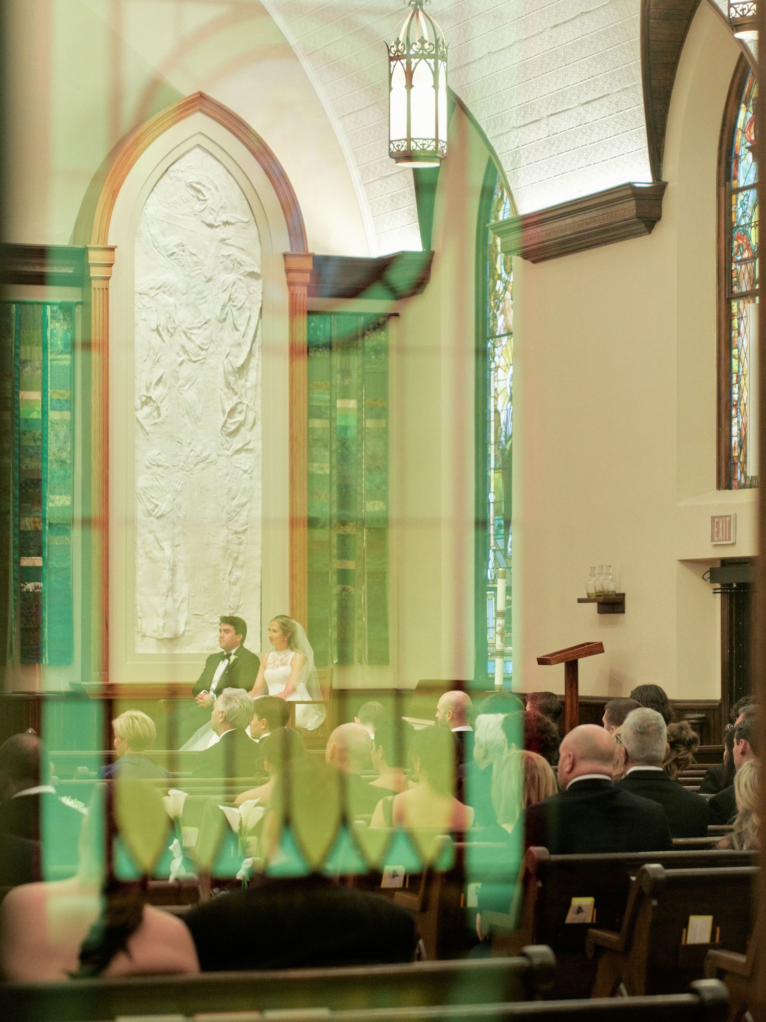 A wedding ceremony inside a church with green stained glass windows, bride and groom at the altar, guests seated in wooden pews.