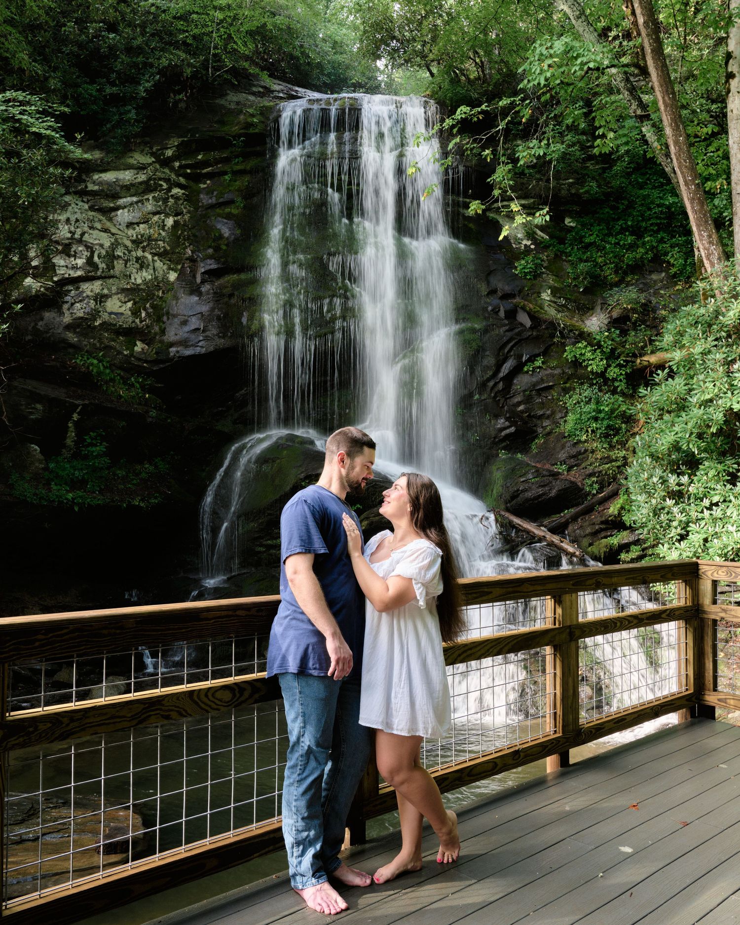 Couple sharing a romantic moment on a wooden deck in front of a stunning waterfall surrounded by lush greenery.