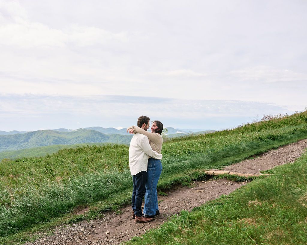 Two people embrace on a hilltop trail with sweeping mountain vistas and green rolling hills in the background.