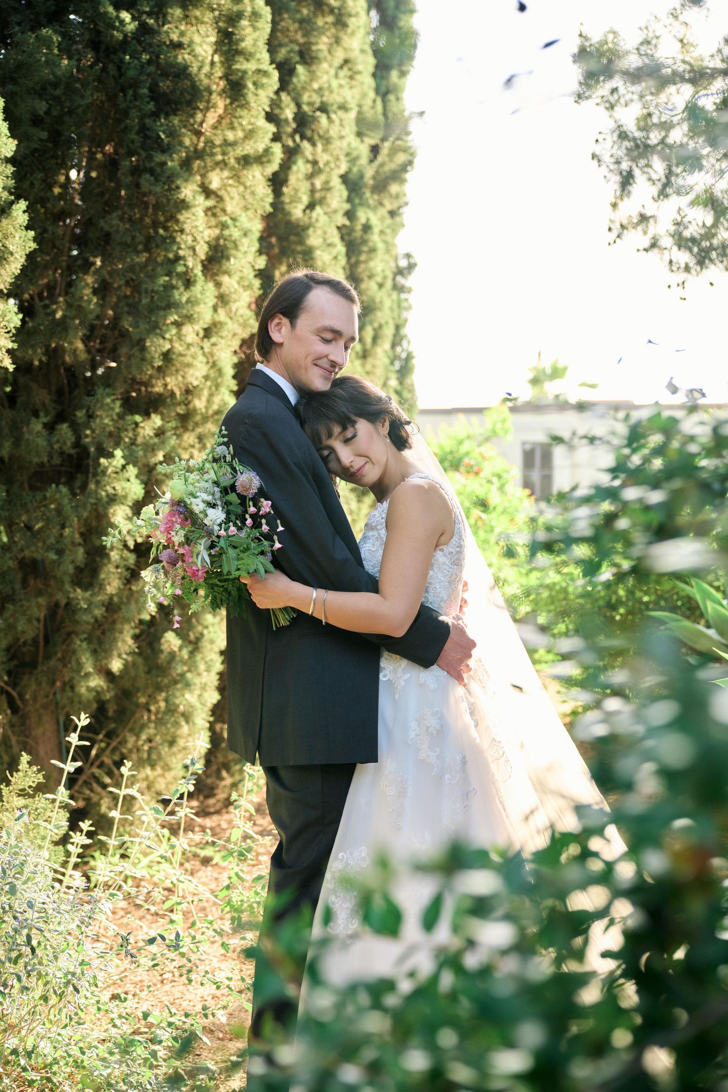 A romantic wedding portrait of a couple embracing among cypress trees in a garden setting.