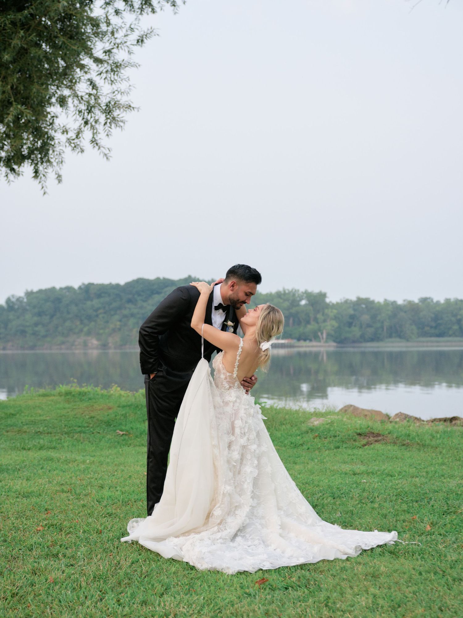 A couple shares a romantic moment by a serene lake, with the bride wearing a flowing lace gown and groom in a black tuxedo.