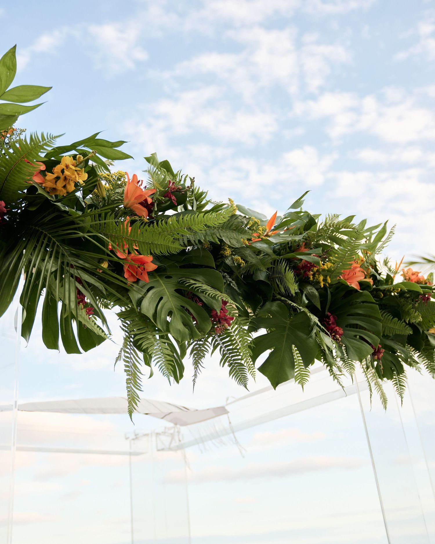 Tropical floral arch arrangement with monstera leaves and orange flowers against blue sky.