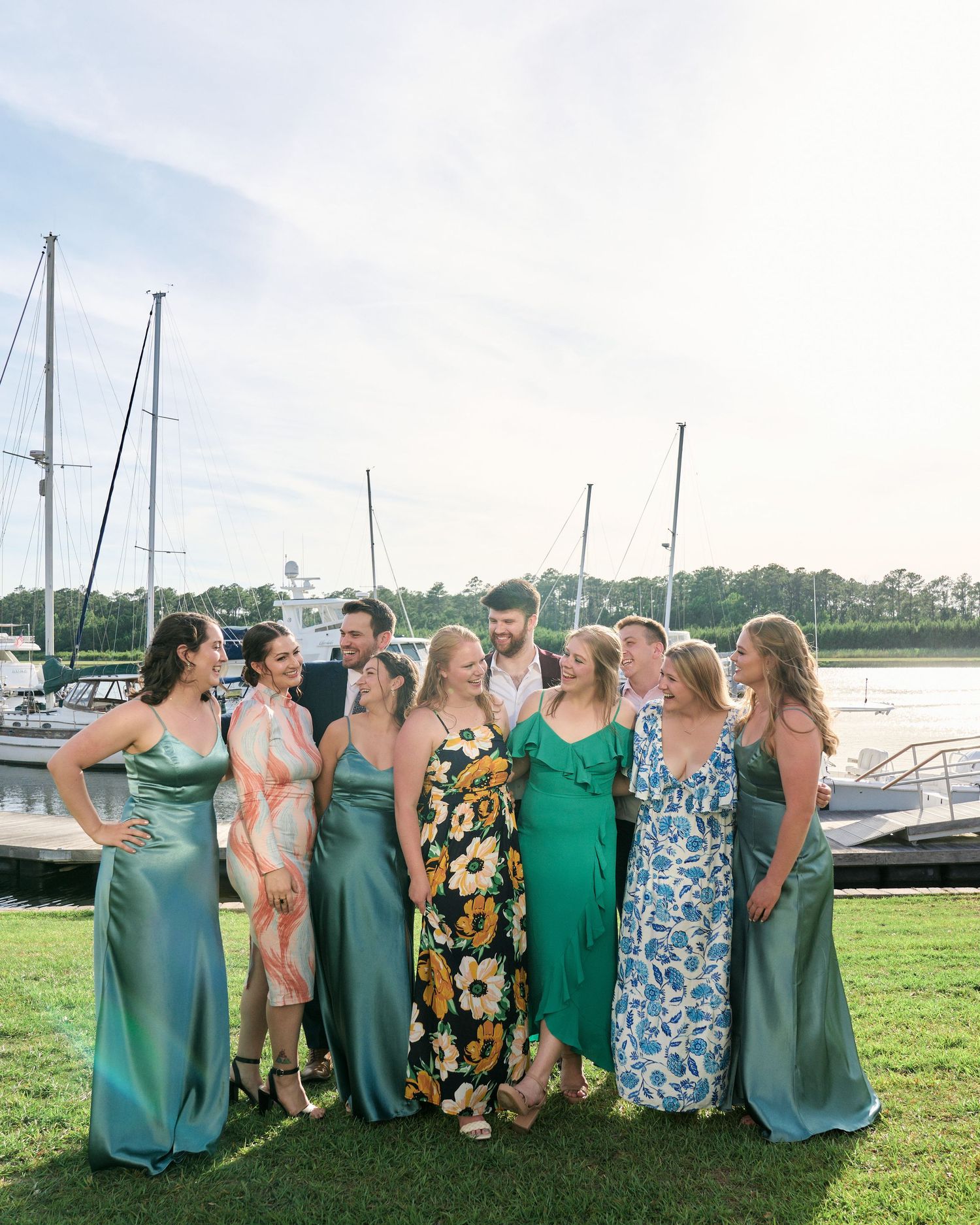 Group in coordinating dresses pose together by a marina with boats and palm trees in the background.