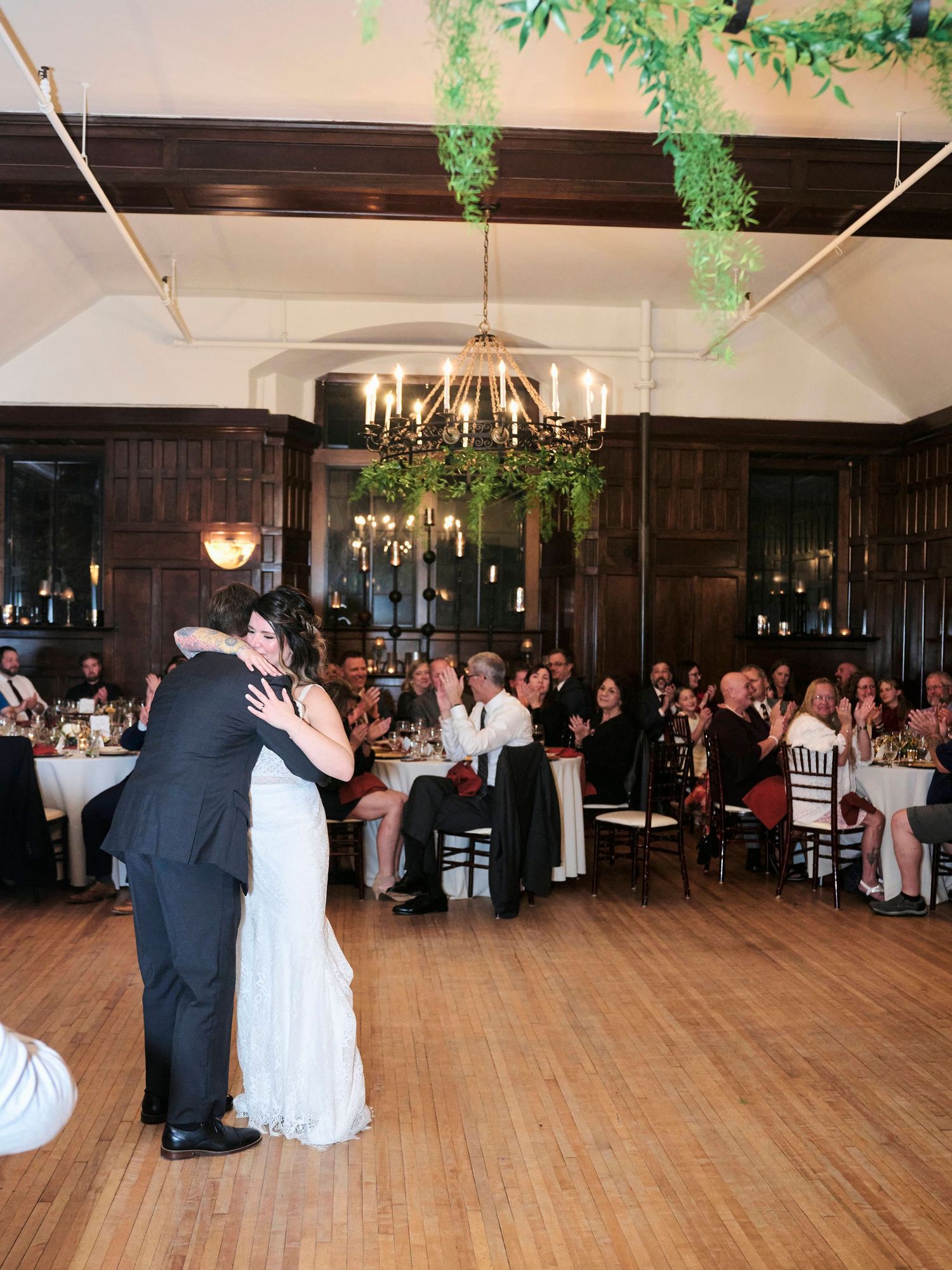 Couple sharing a romantic dance under hanging greenery and chandelier in wood-paneled reception hall.