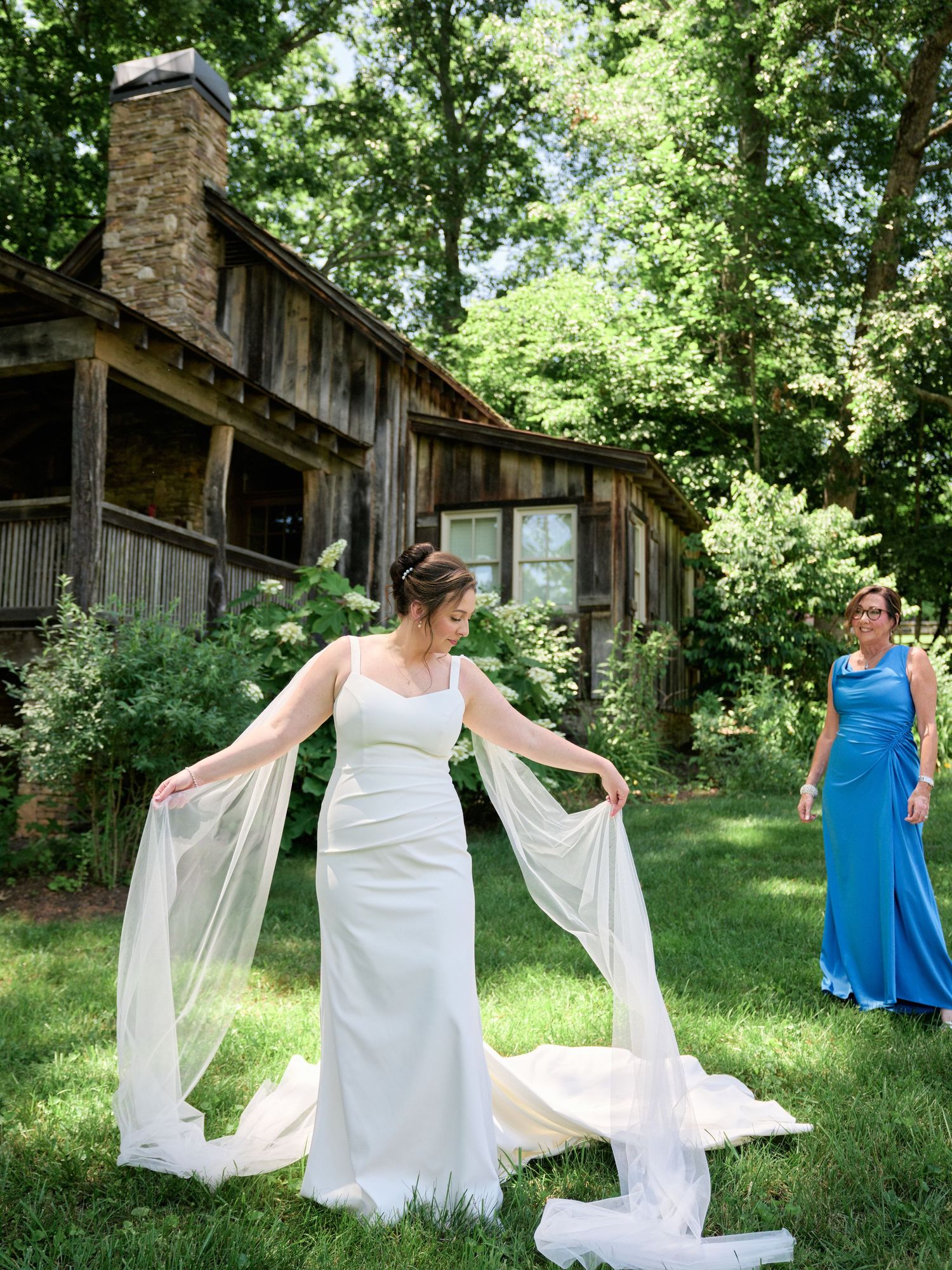 A person in a flowing white wedding dress poses while another in blue looks on by a rustic cabin.