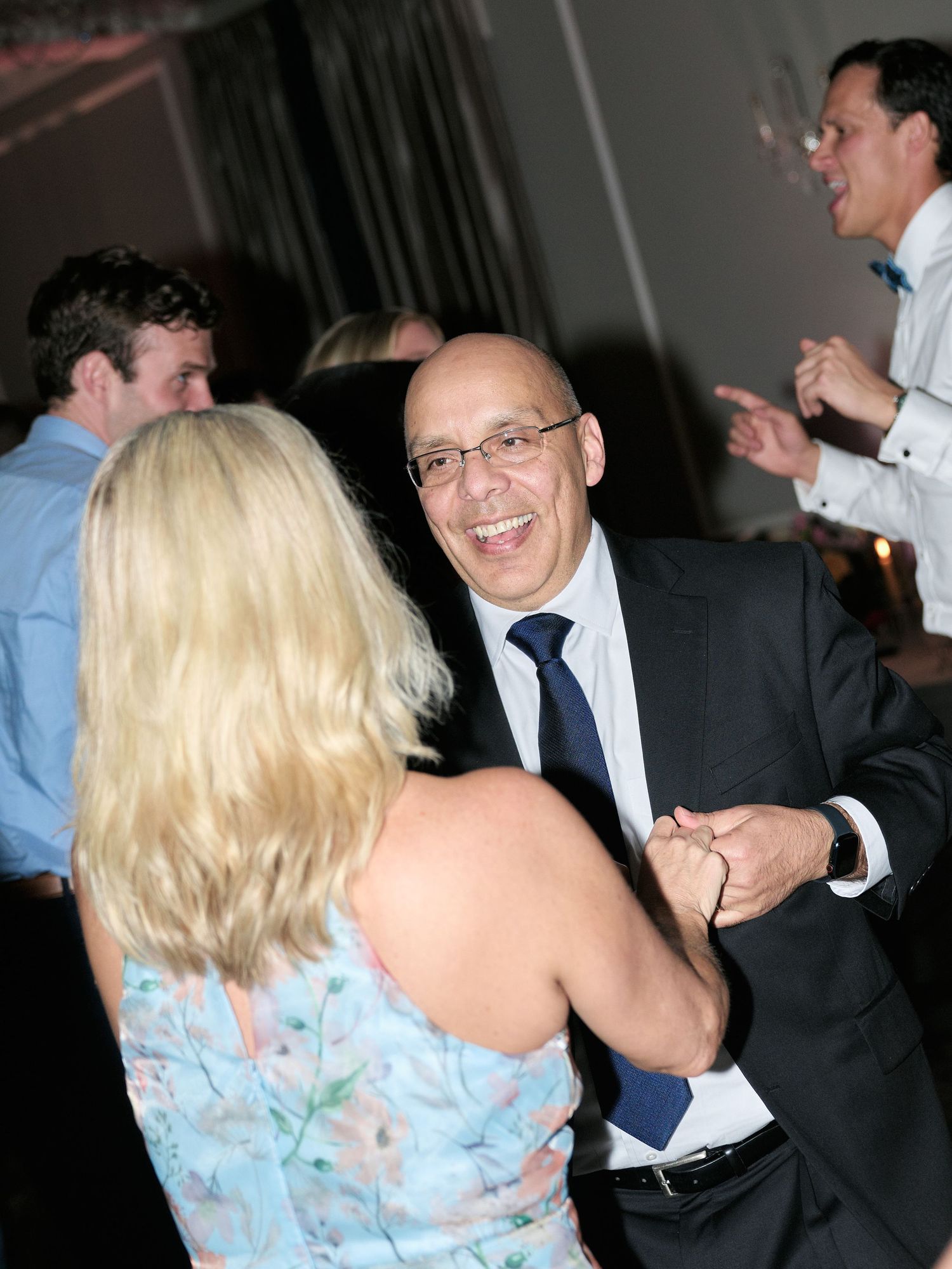 People dancing and laughing at a formal event with a man in suit and glasses on the dance floor.
