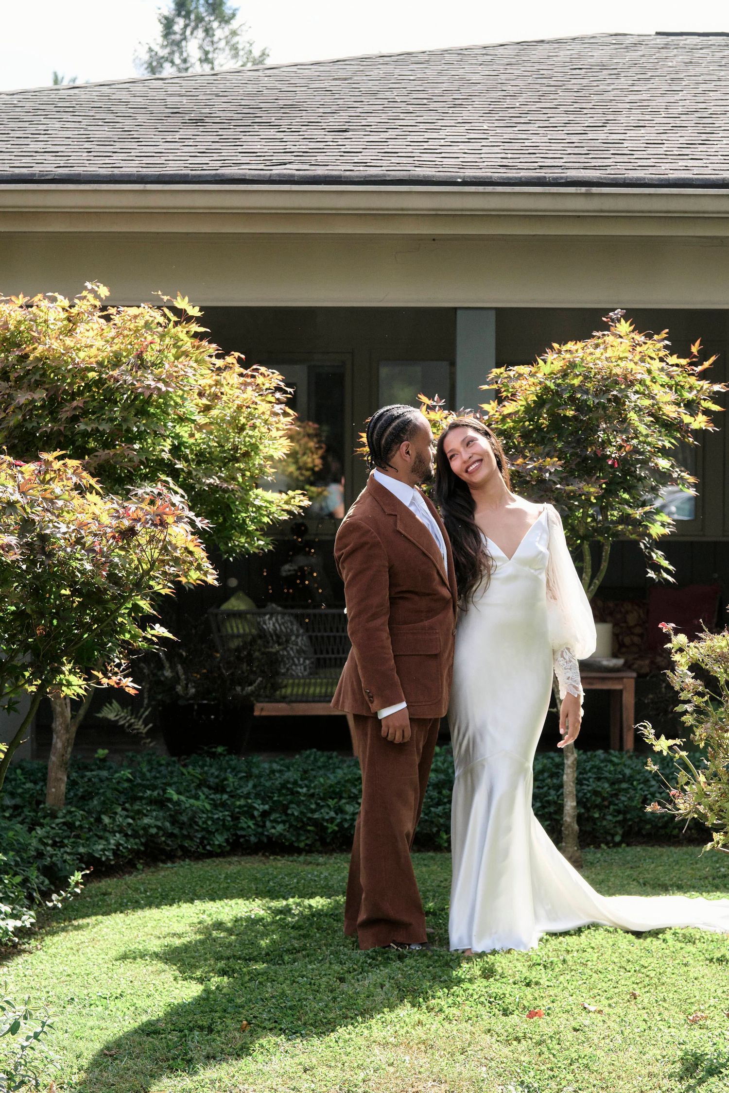 A couple in wedding attire poses together in front of flowering bushes and a house.