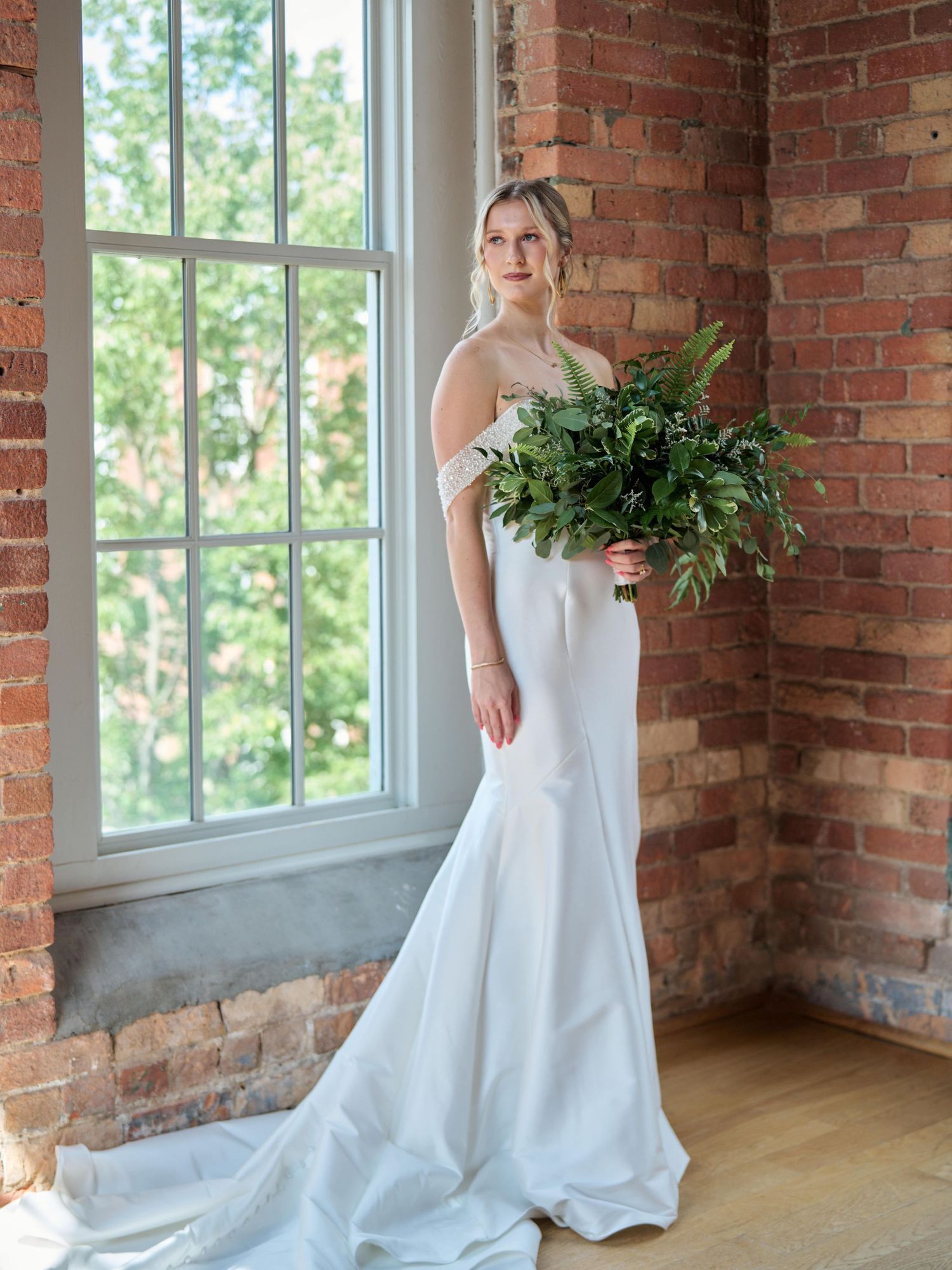 Bride in sleek white wedding dress holds greenery bouquet by brick window in natural light.