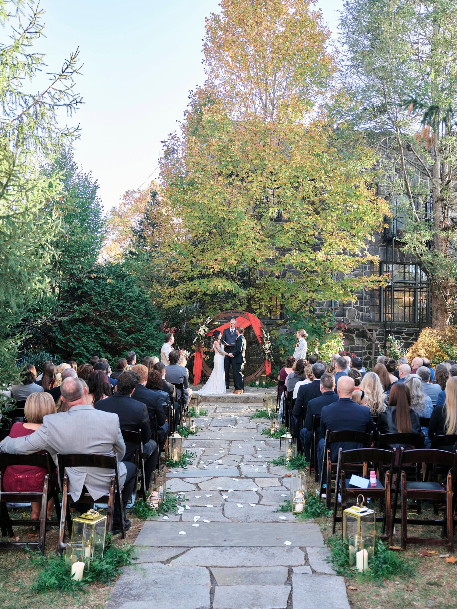 Outdoor wedding ceremony with guests seated on either side of a stone path leading to a red circular arch.
