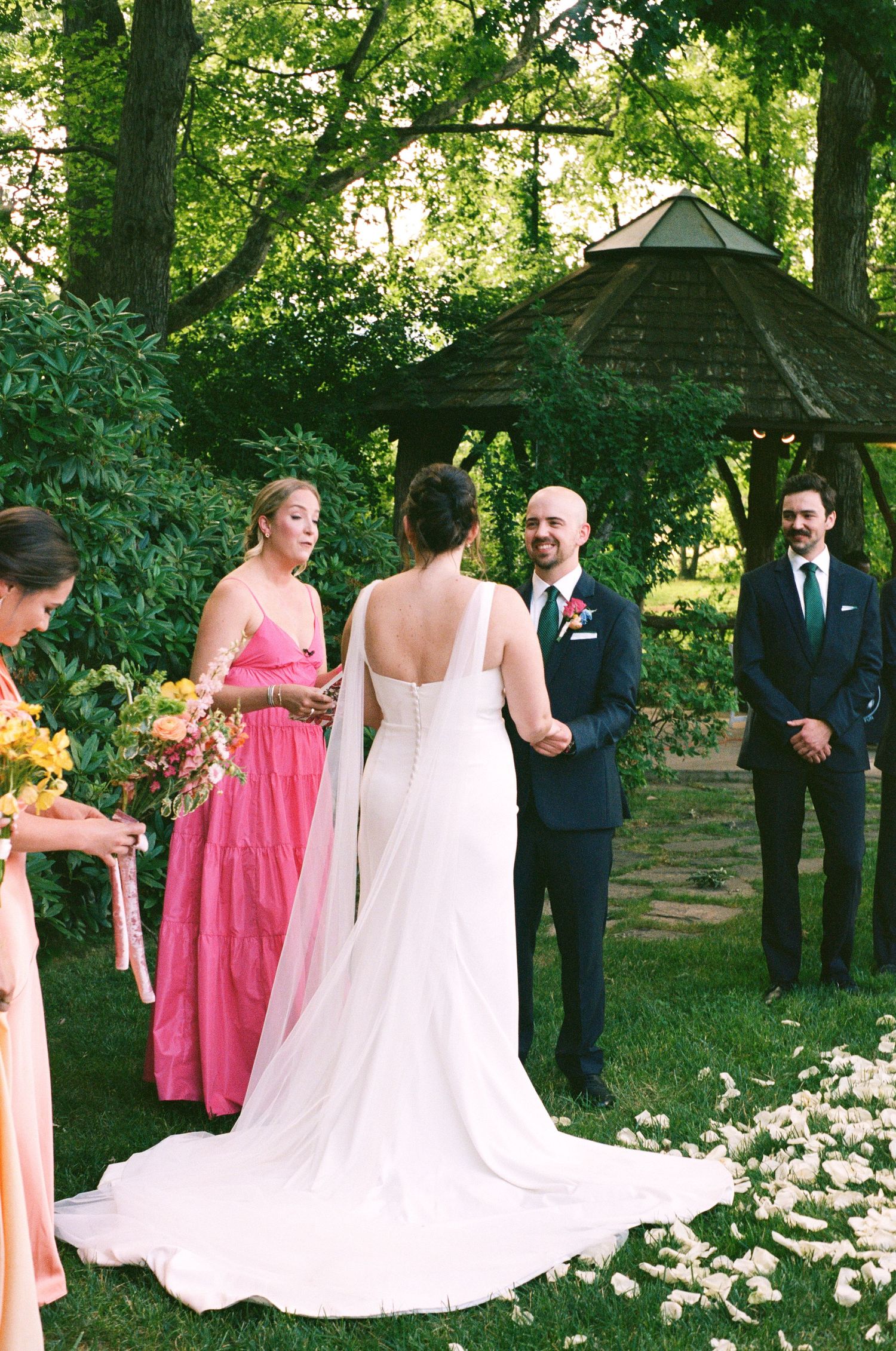 Outdoor garden wedding ceremony with pink bridesmaid dresses and flower petals lining the aisle.