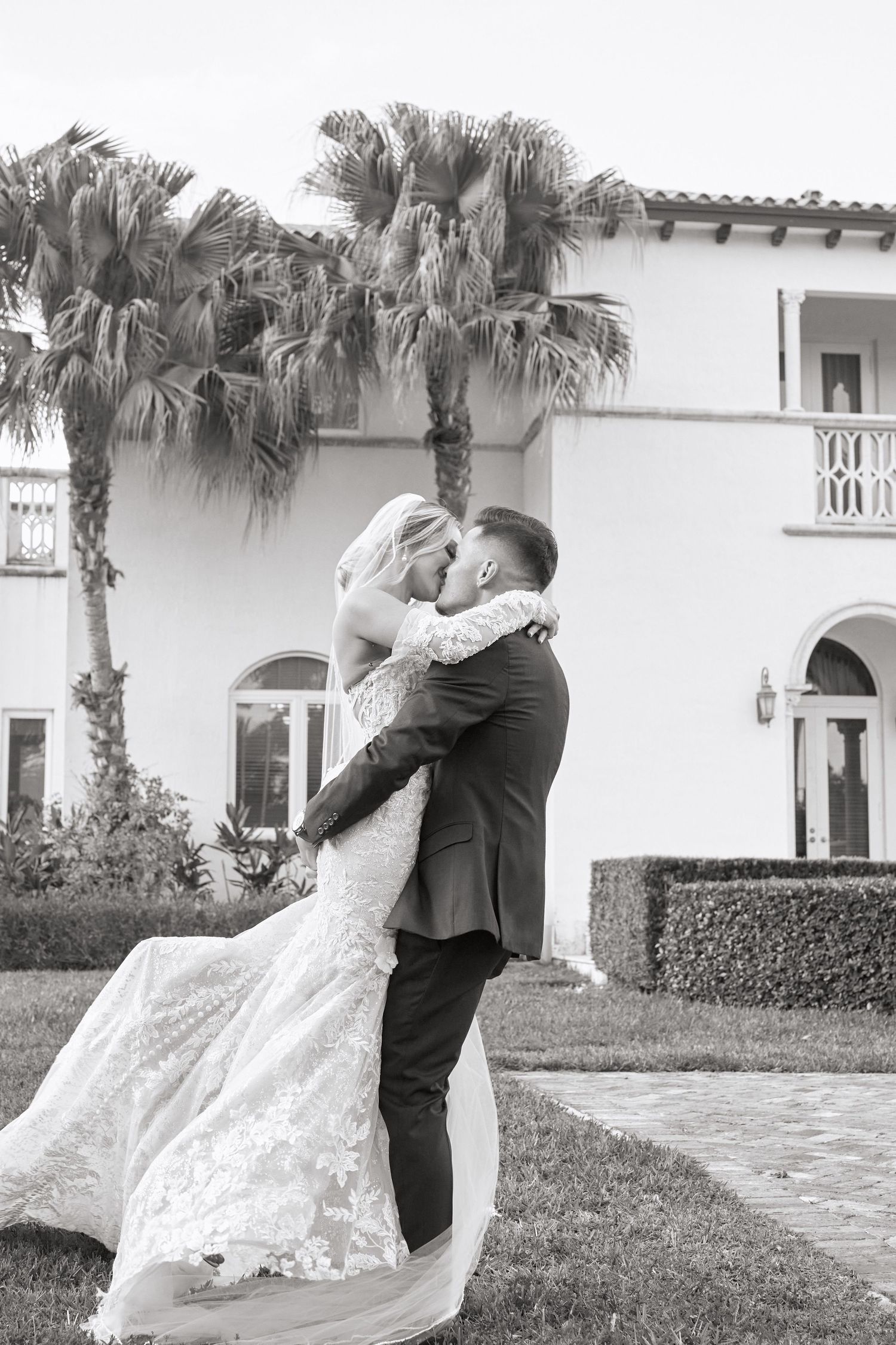 A romantic wedding couple embraces in front of a Mediterranean-style villa with palm trees.