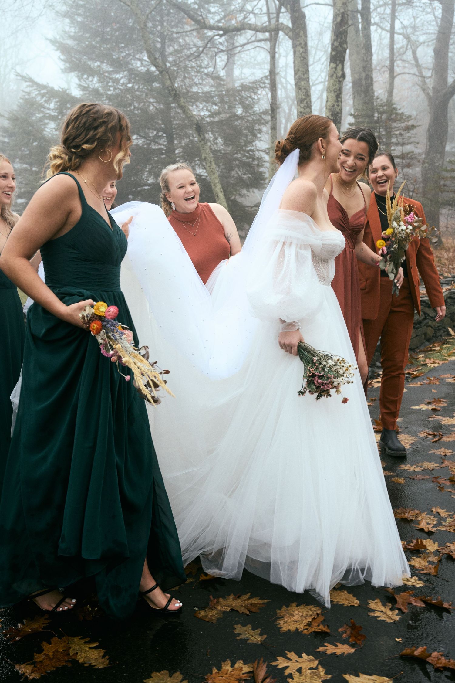 A bride in a white gown laughs with bridesmaids wearing jewel-toned dresses on a foggy autumn day.